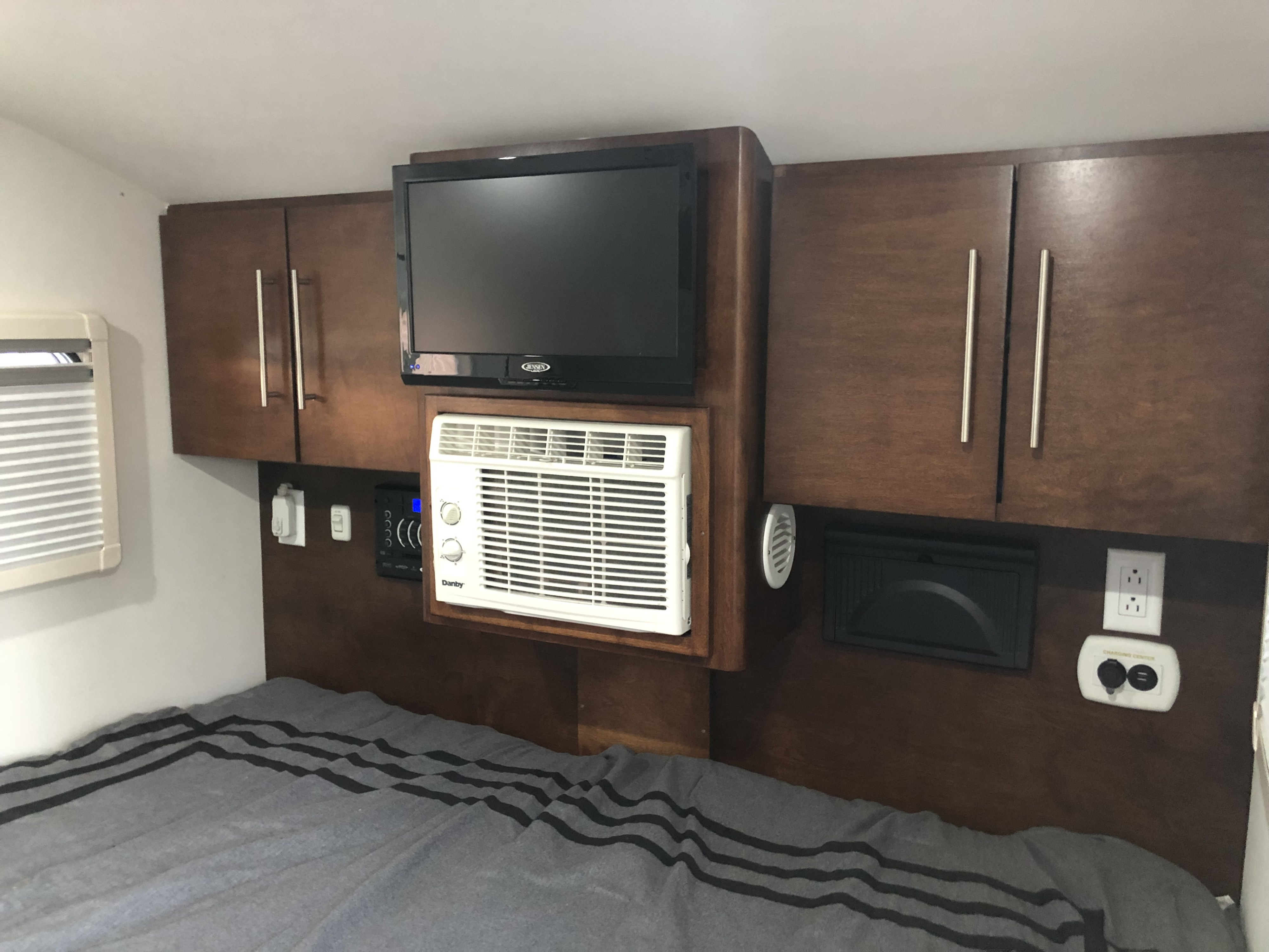 Cozy RV rental bedroom featuring a wall-mounted TV, compact AC unit, cabinets, and a charging center above a striped bed.. T@G XL 2017