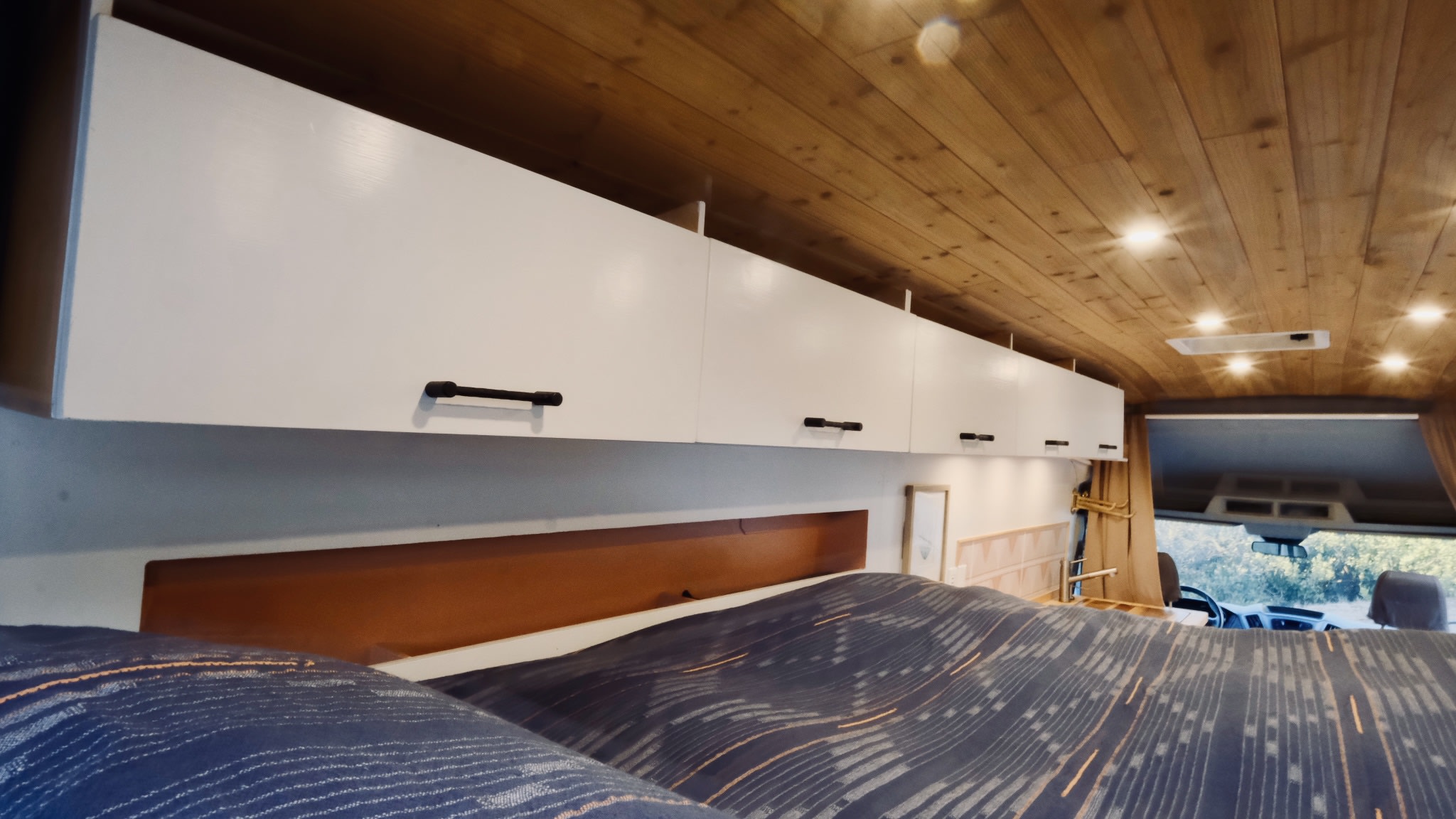 View from the bed in this RV rental van. It features white overhead cabinets, a wooden ceiling with lights, and front cabin views.. Ford Transit 2016