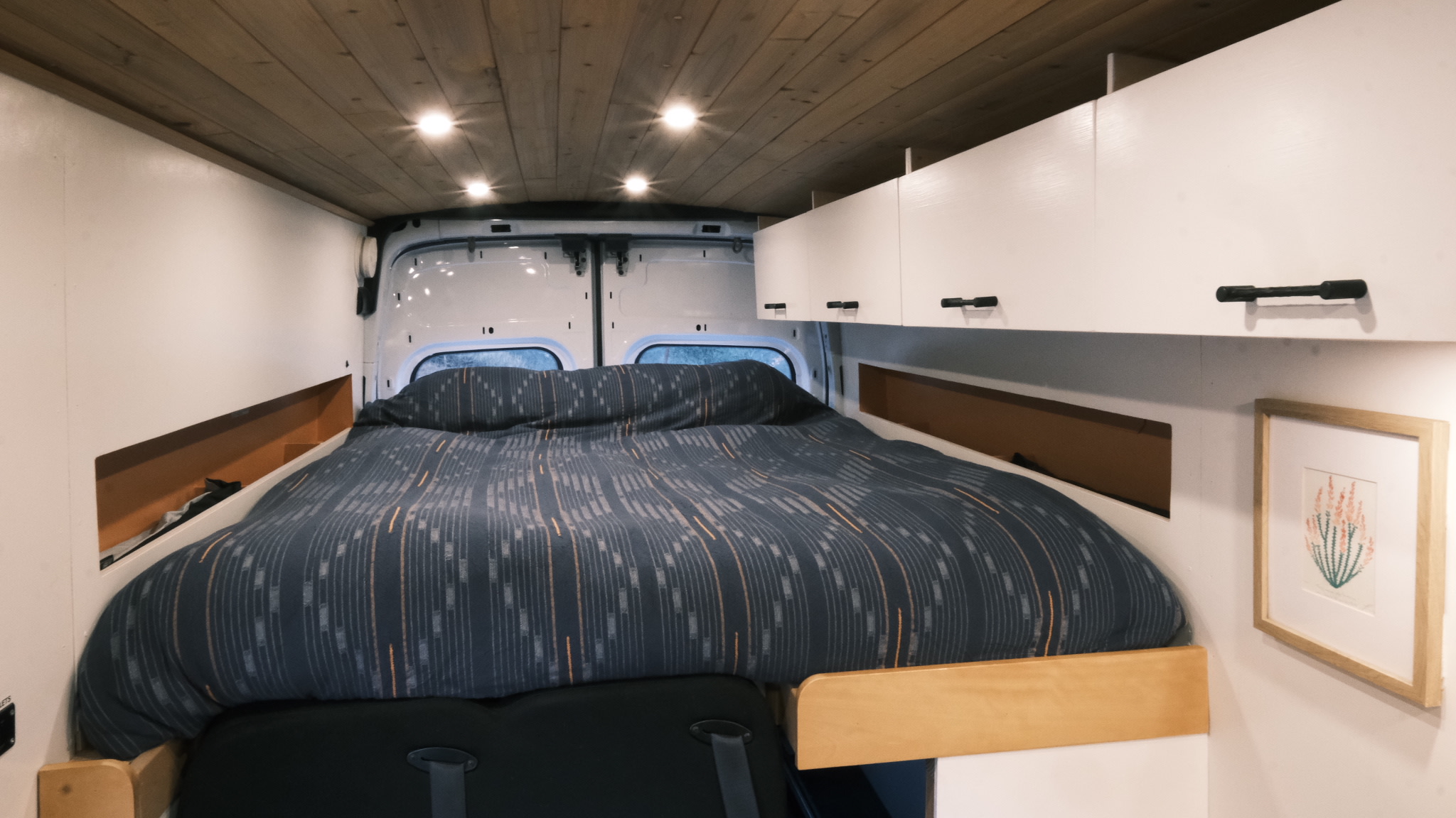 RV rental interior: cozy camper van bedroom with patterned duvet, overhead cabinets, and a wood-paneled ceiling.. Ford Transit 2016