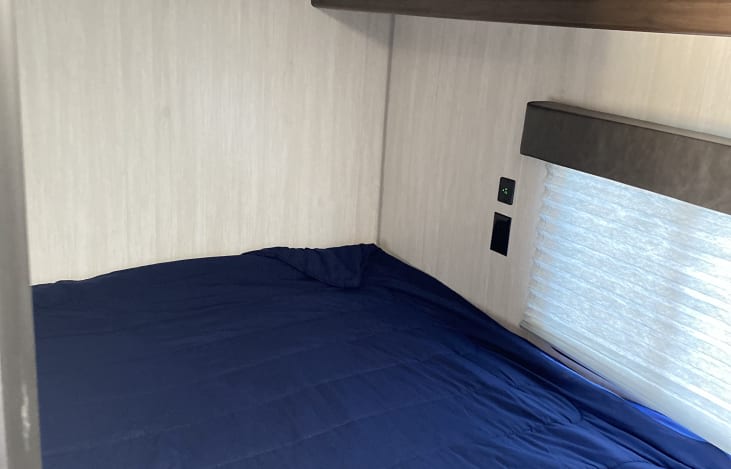 RV rental bedroom featuring a bed with dark blue bedding, light paneled walls, window blind, and power outlets.. Forest River Cherokee Grey Wolf 2020