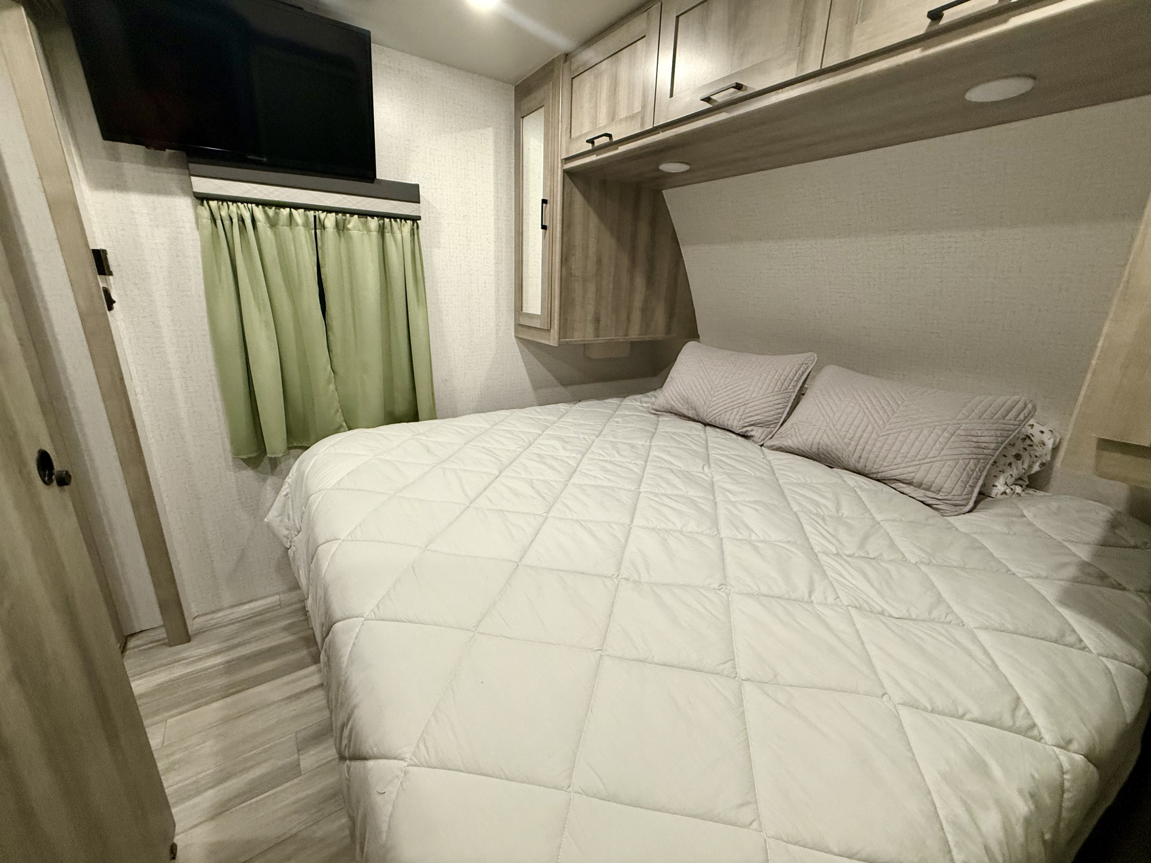 RV rental bedroom with a quilted bed, overhead cabinets, TV, and green curtains, providing a cozy and functional space.. Winnebago Voyage 2022