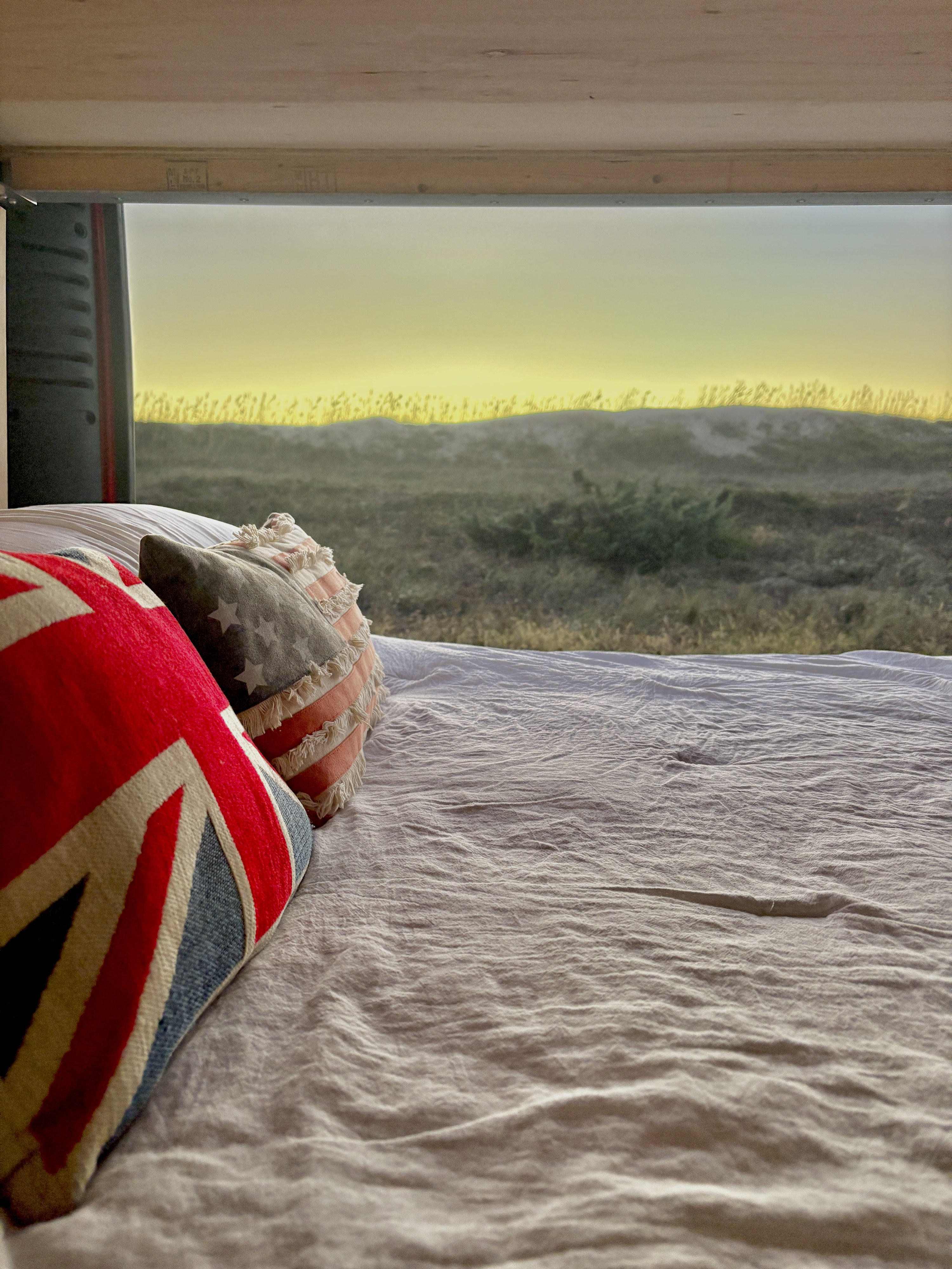 RV rental bed with patriotic pillows (Union Jack, US flag) offering a serene view of dunes under a soft yellow sky.. Ram Promaster 3500 2019