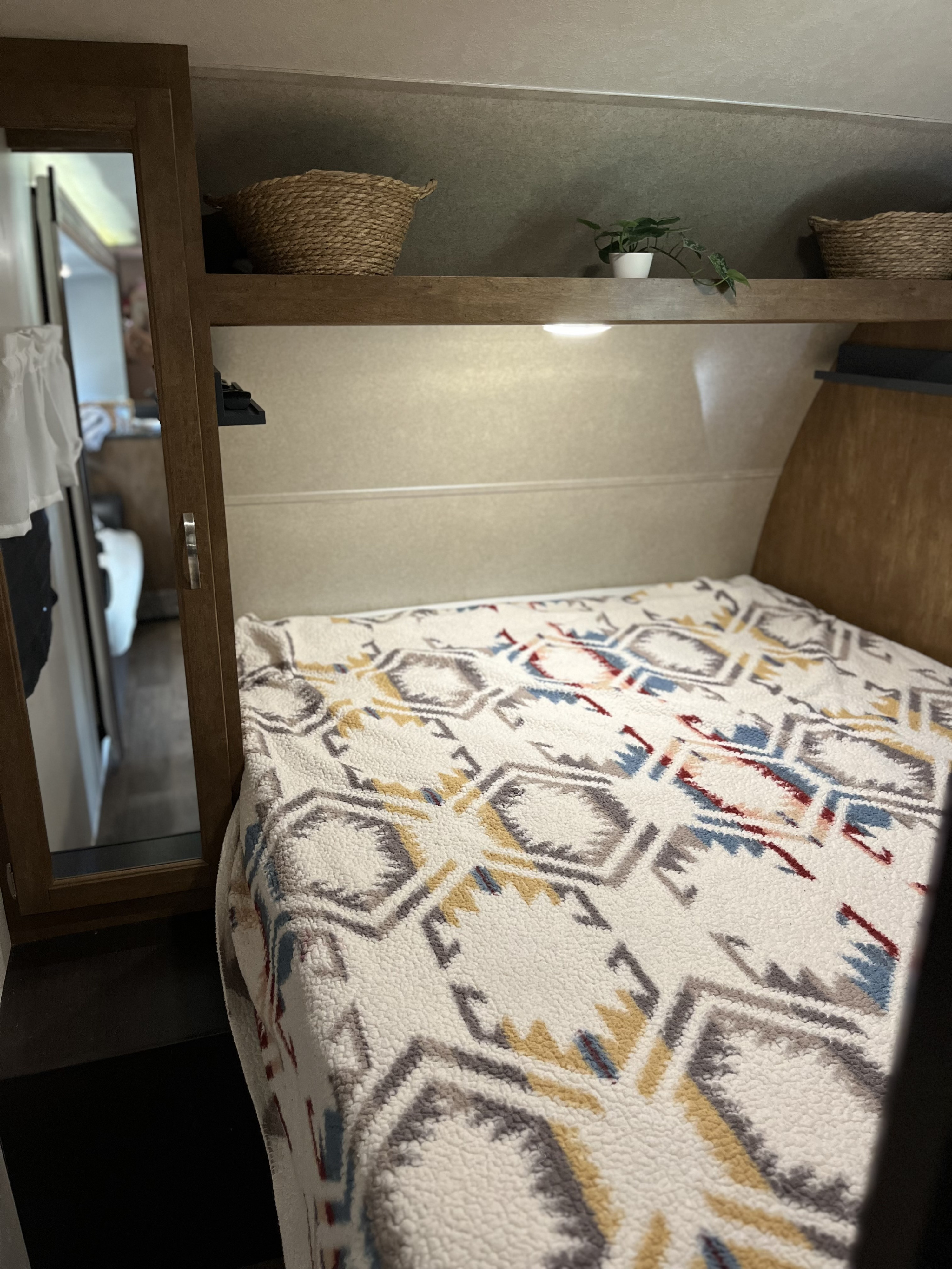 Inside an RV rental: a compact sleeping area features a bed with a patterned blanket, an overhead shelf with baskets, and a small plant.. Forest River Wildwood X-Lite 2018