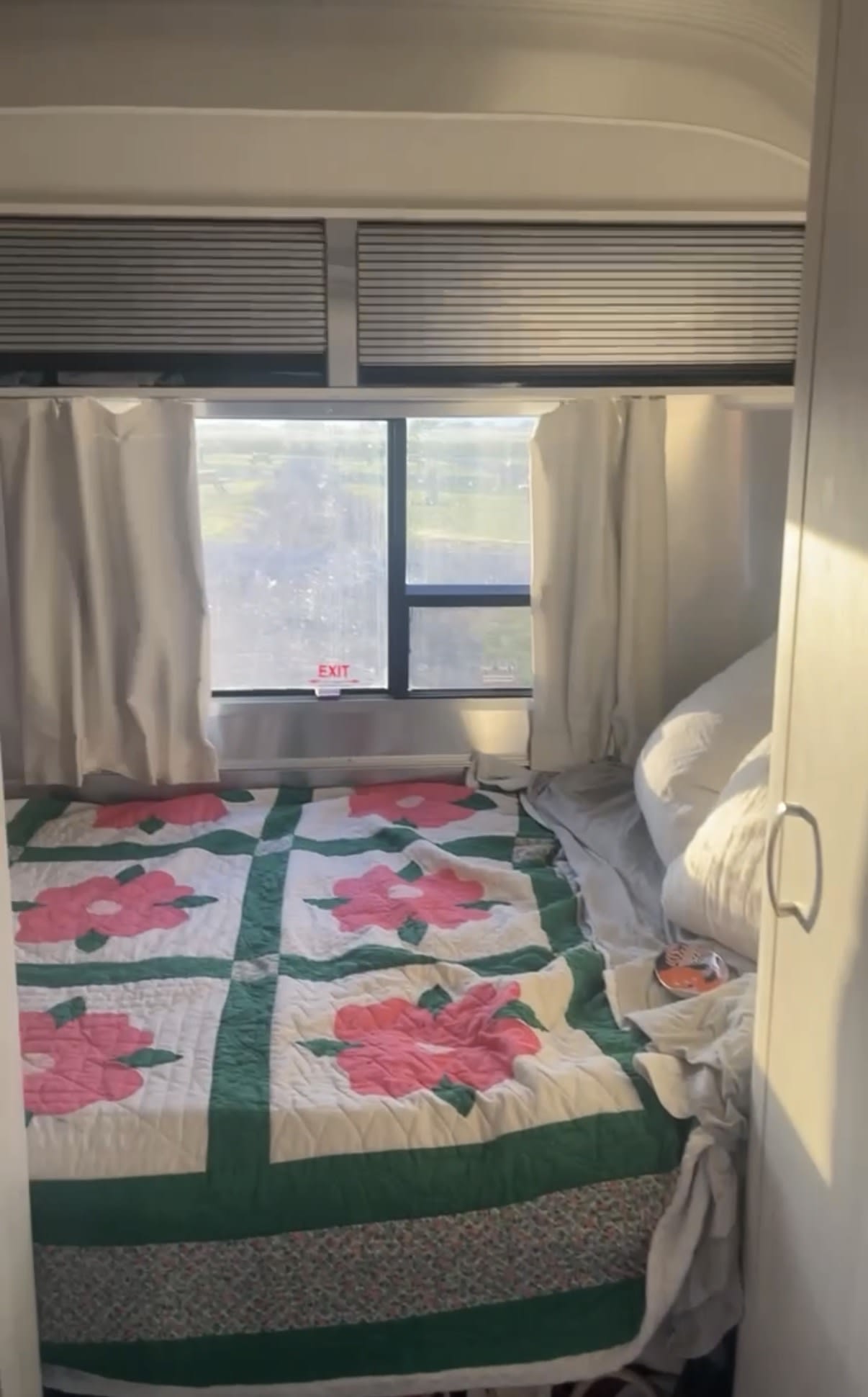 Inside an **rv rental**, a bed with a colorful floral quilt and white pillows sits by a window with an 'EXIT' sign, offering a cozy sleep space.. Airstream Sport 2019