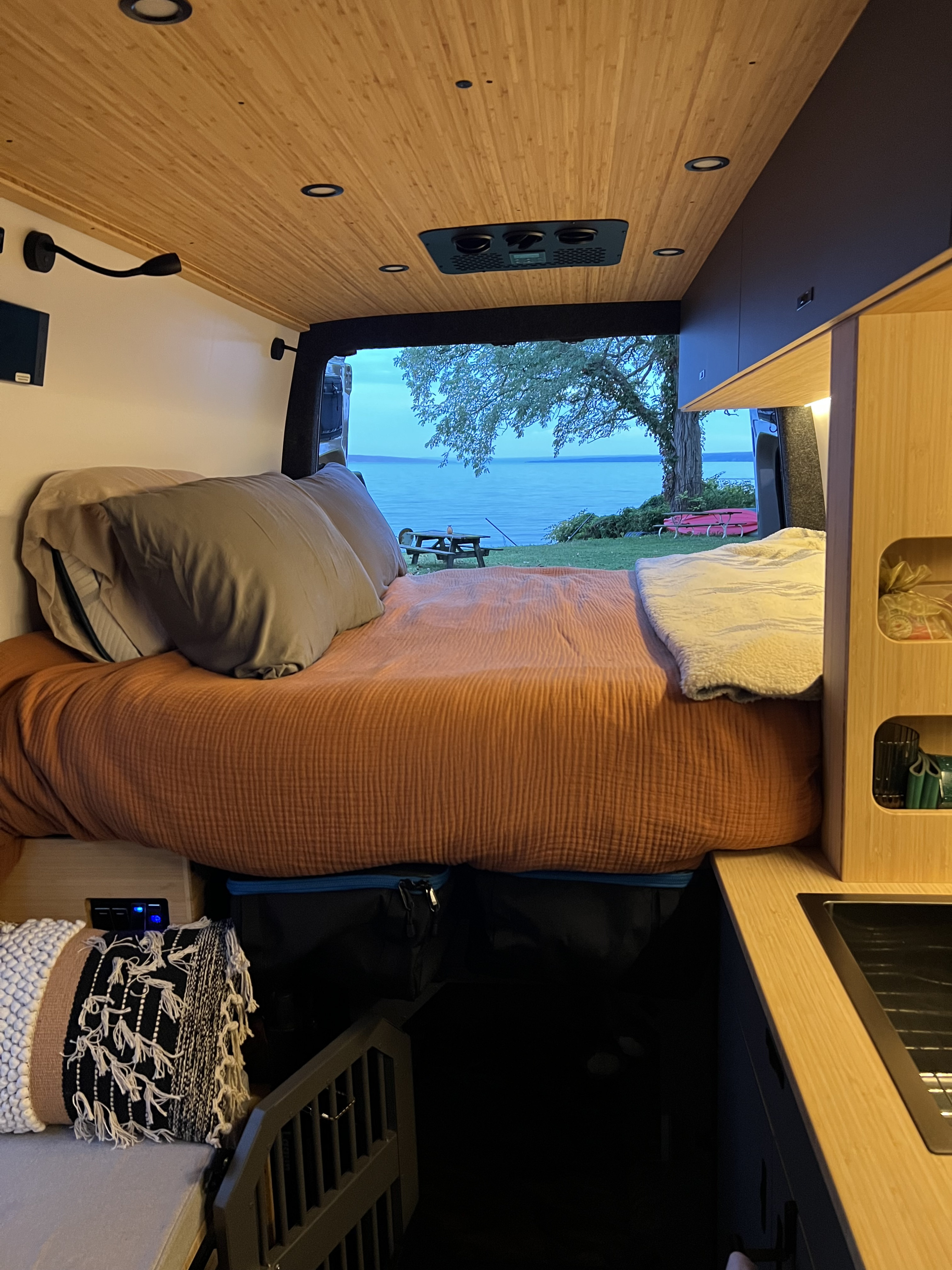 Cozy rv rental interior featuring a comfy bed, modern wooden ceiling, and a stunning lake view from the open rear doors.. Mercedes Sprinter 2025