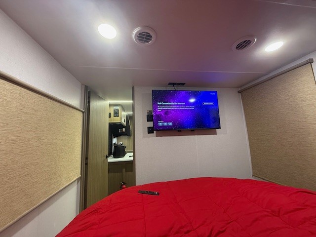 Inside this rv rental: comfy bedroom with red comforter, mounted TV (internet disconnected), and kitchen access.. Forest River Wildwood 2025