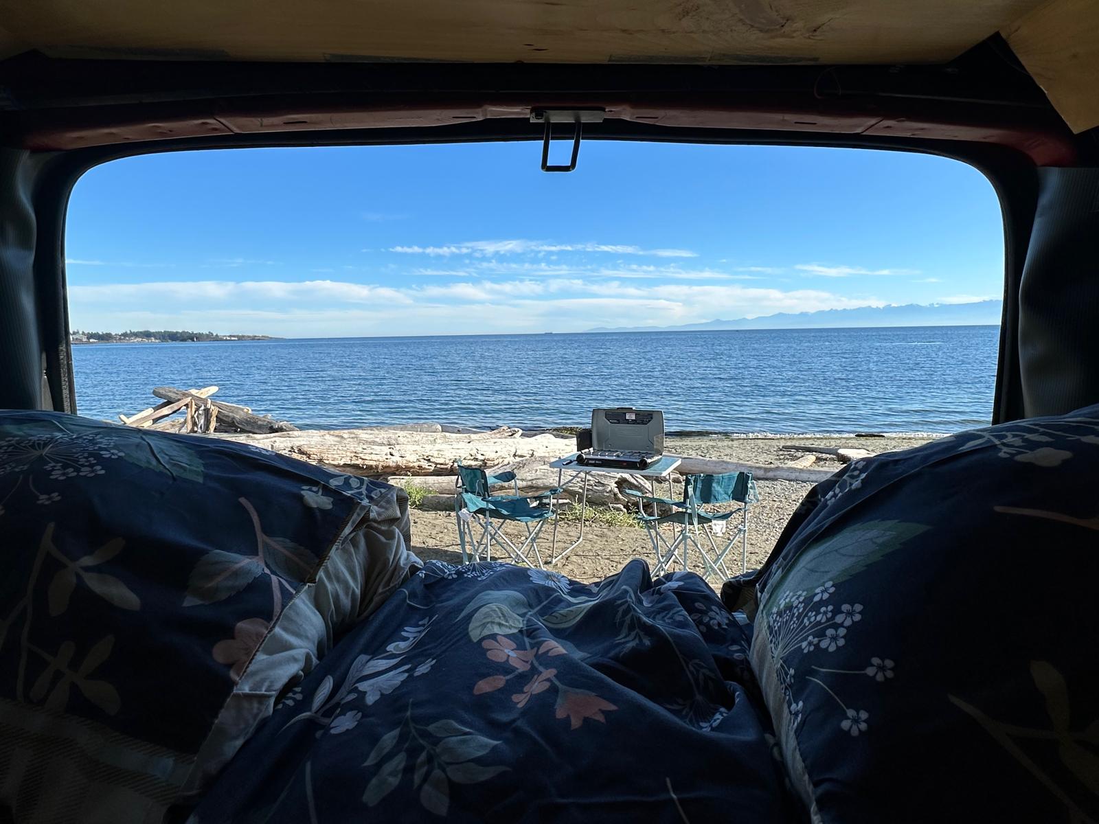 Cozy rv rental view: Floral bedding inside, looking out at a serene beach with driftwood, a portable stove, and blue ocean.. Gmc Savanna 2003