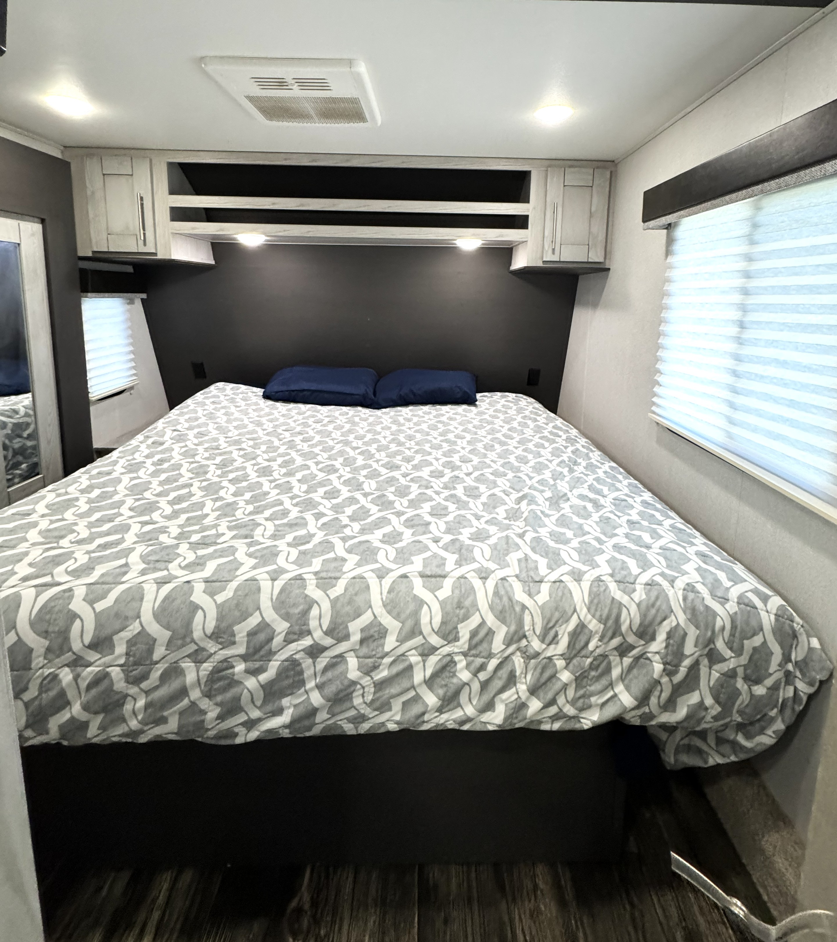 A cozy RV rental bedroom features a patterned bed, blue pillows, overhead storage, and window blinds.. Keystone Fuzion 2021