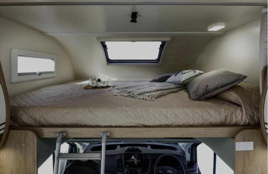RV rental's over-cab sleeping area: a neatly made bed with pillows, blanket, windows, and dashboard visible below.. RollerTeam Auto Roller 746 2026