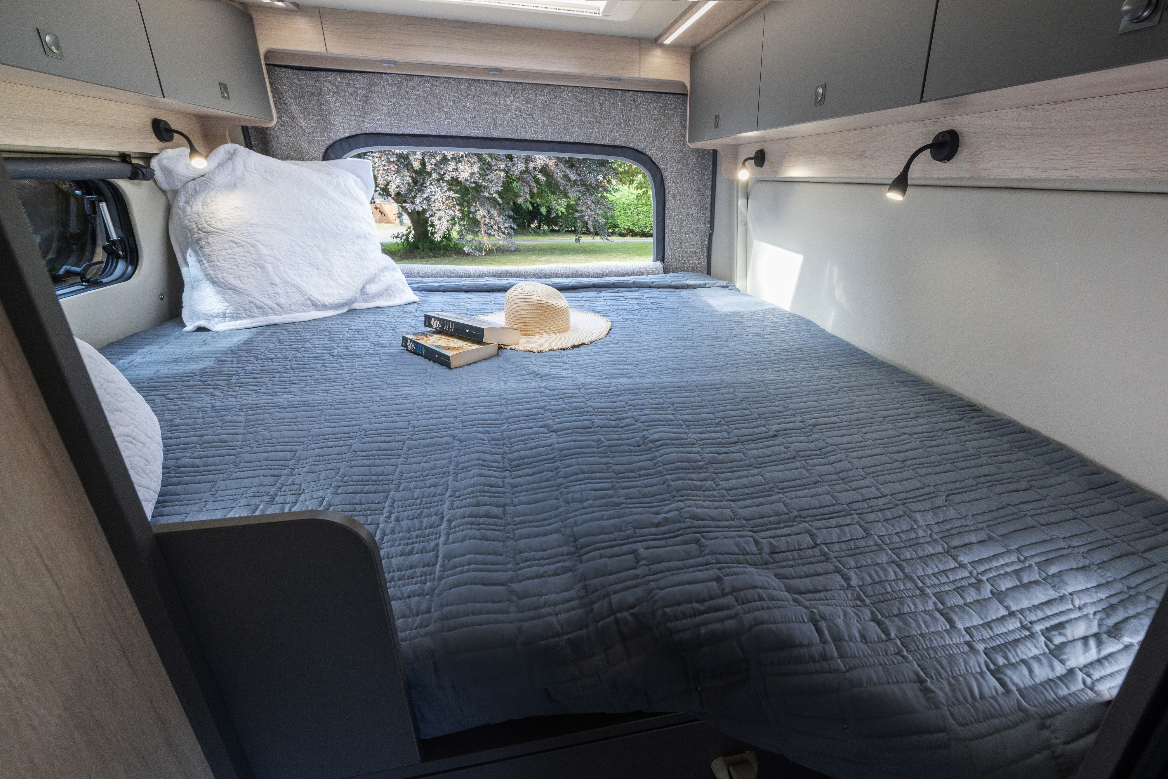 Cozy RV rental bedroom with a comfortable bed, reading lights, and a serene window view of nature. Books and hat await.. Auto Trail Expedition 68 XL 2026