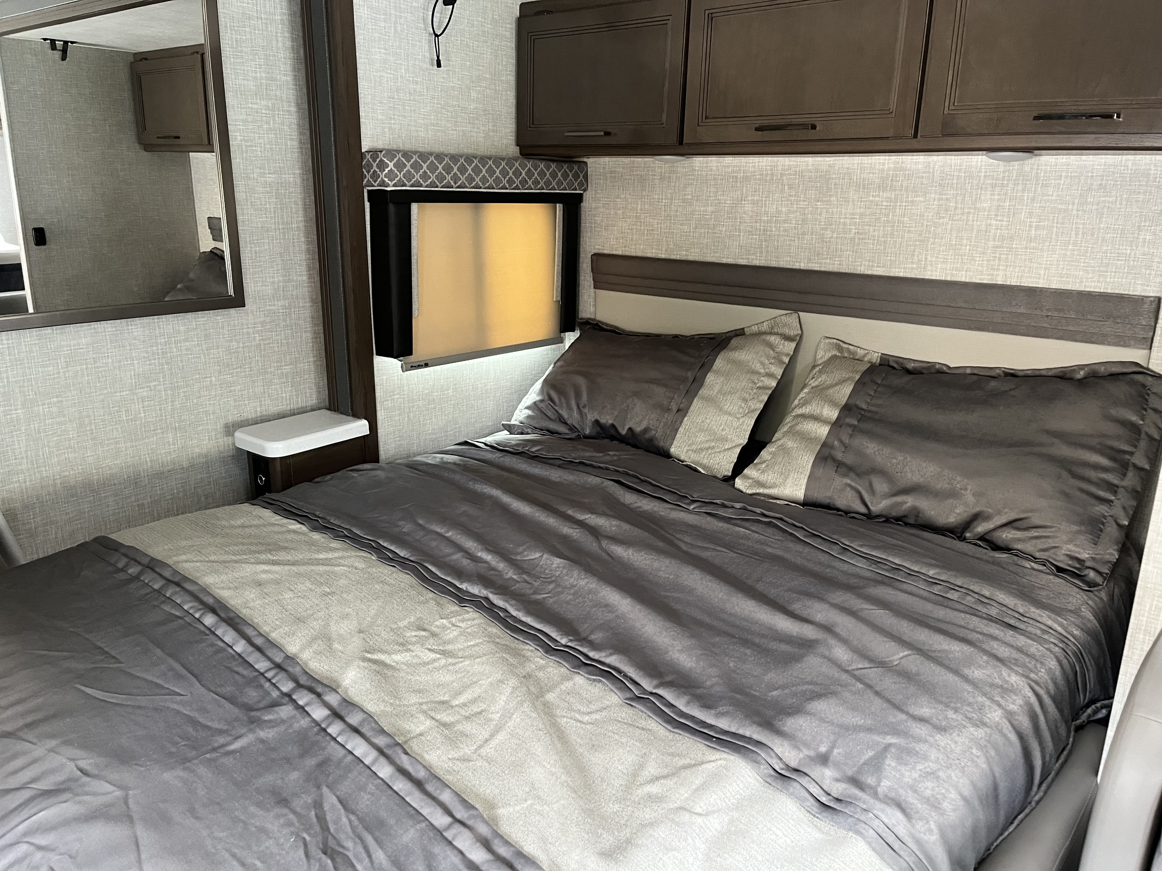 This RV rental bedroom has a bed with gray & light gray bedding, overhead cabinets, a window, and a mirror.. Thor Quantum 2022