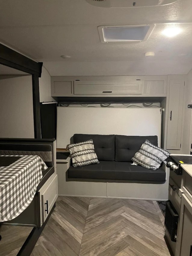 Cozy RV rental interior with a gray sofa, patterned pillows, bunk bed with checkered linen, and modern herringbone floor.. Forest River Wildwood FSX 2021