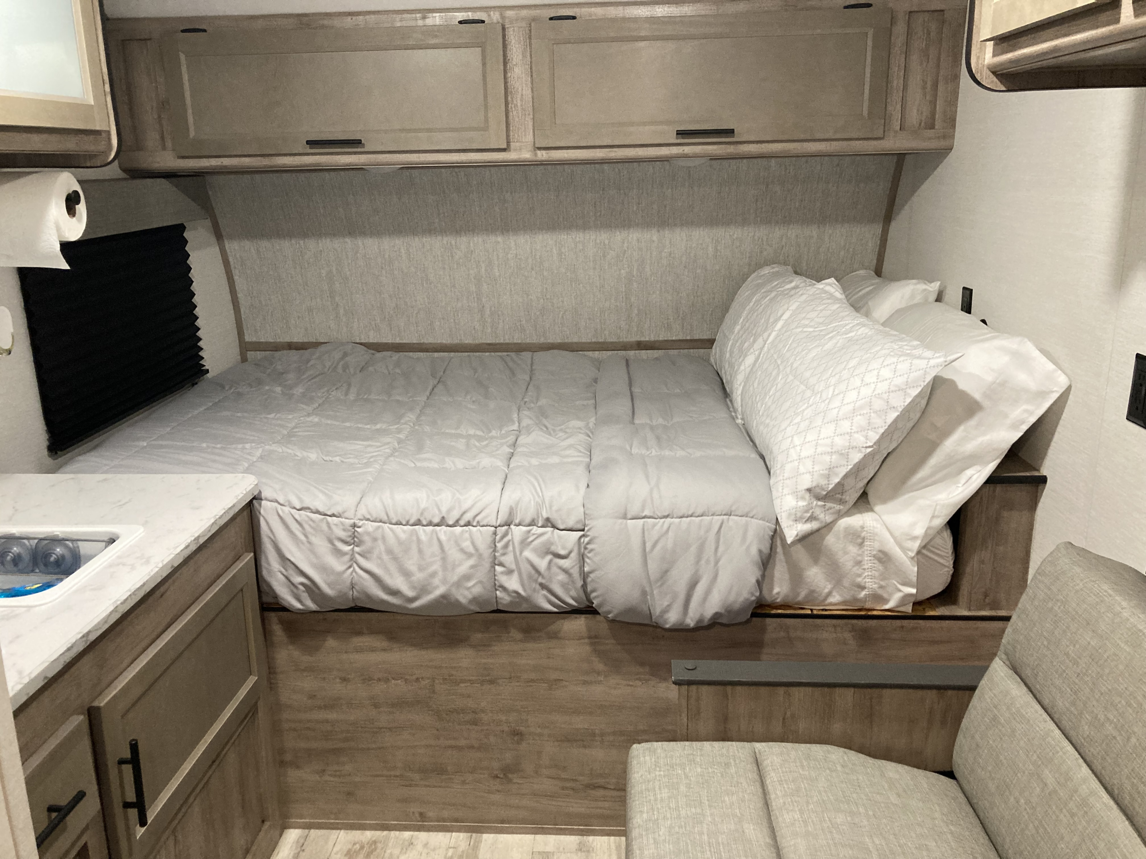 Cozy RV rental interior featuring a bed with light gray bedding, overhead cabinets, a small sink, and a cushioned seat.. Gulf Stream Yellowstone Trail Edition 150RD 2025