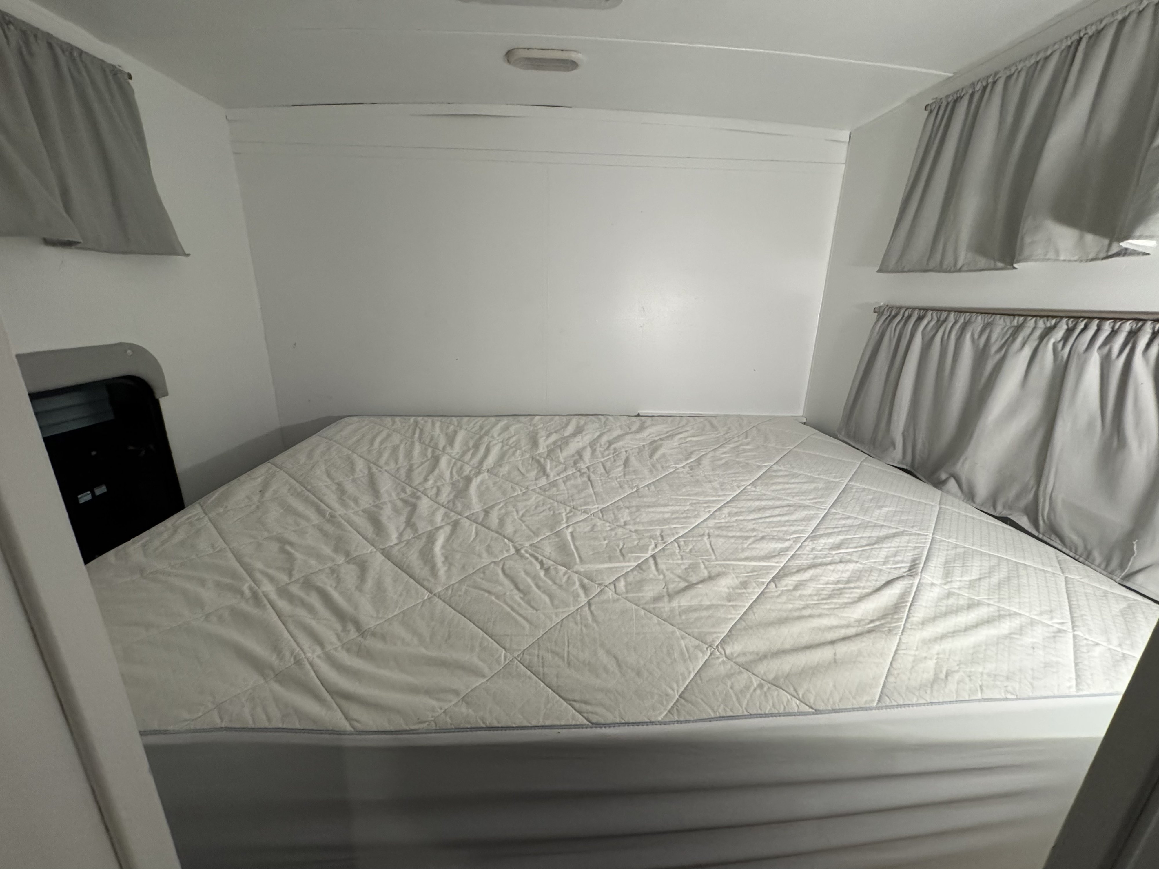 Inside a bright rv rental bedroom, a white quilted mattress pad covers the bed, complemented by simple gray curtains.. Forest River Rockwood Mini Lite 2015
