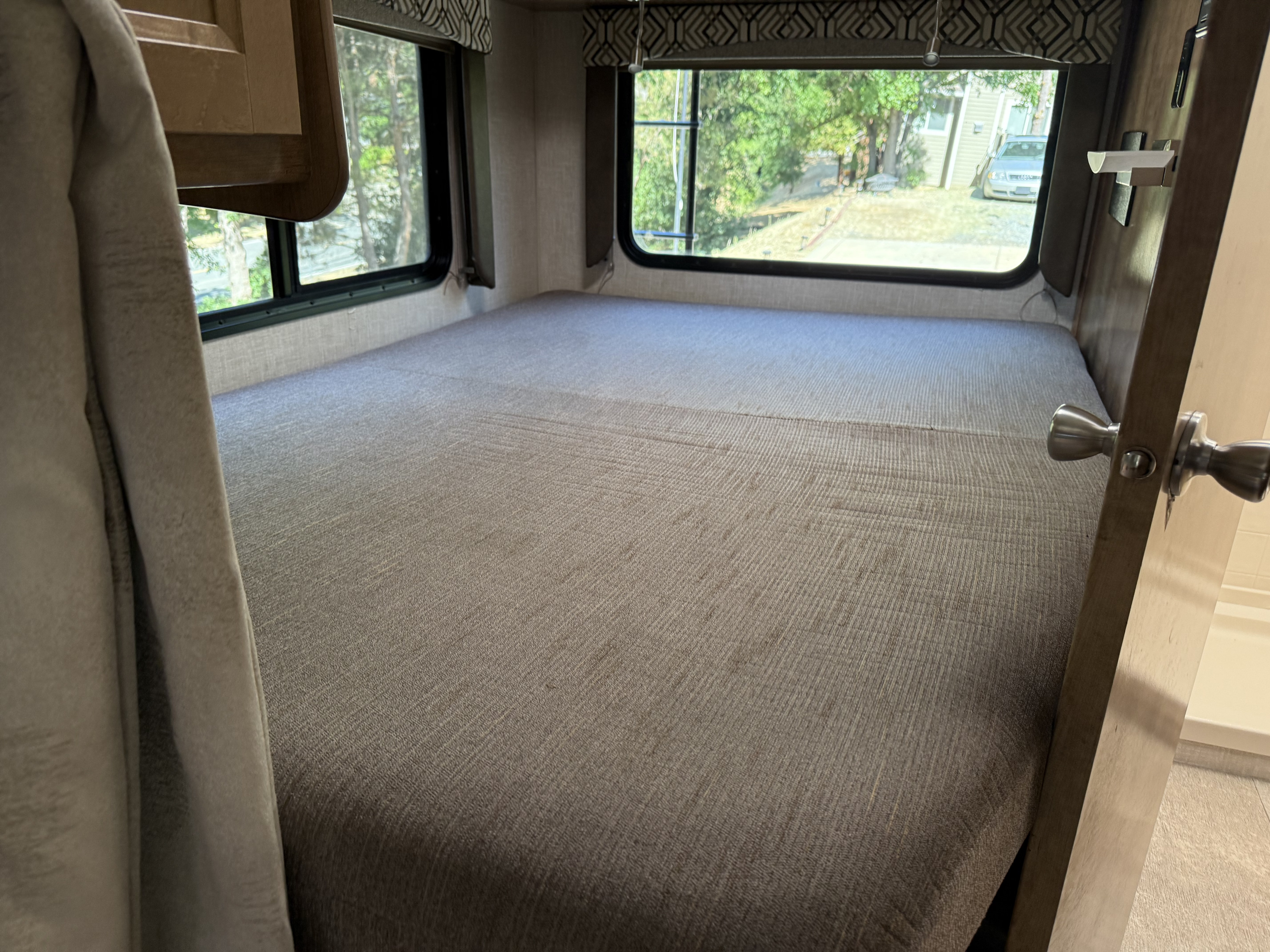 RV rental bedroom: a large bed fills the space, flanked by windows showing trees, with a partial door visible on the right.. Forest River Coachmen Leprechaun 2020