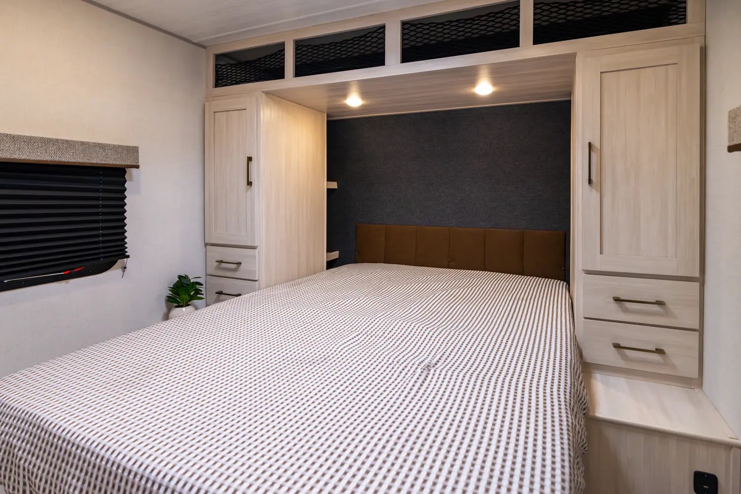 Inside a cozy RV rental bedroom: bed with striped blanket, flanked by light wood cabinets, overhead storage, and window.. Forest River Surveyor X 27CAMP 2025