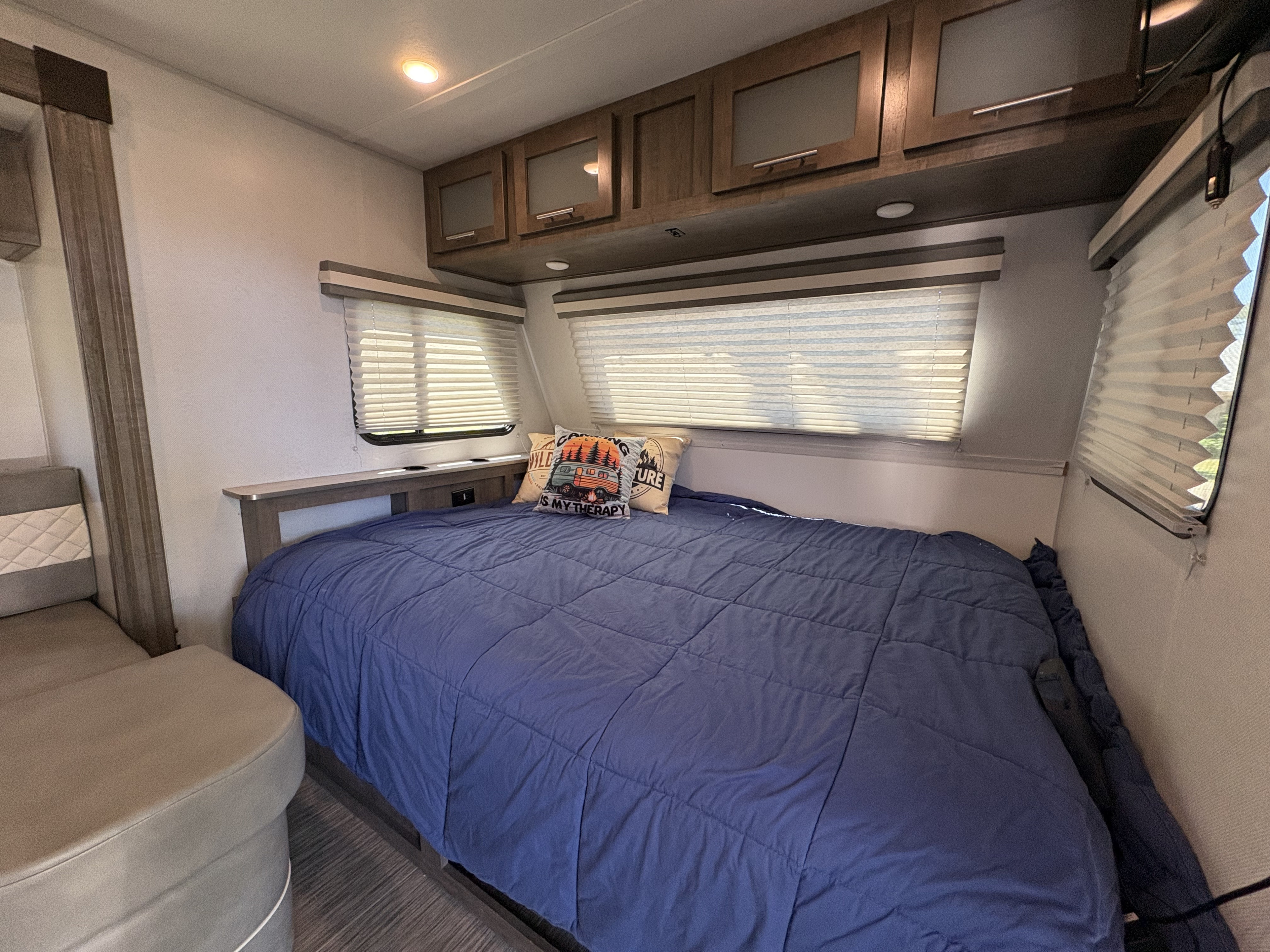 This RV rental offers a cozy sleeping area with a blue bed, camping-themed pillows, overhead cabinets, and window blinds.. No Boundaries 19 2020