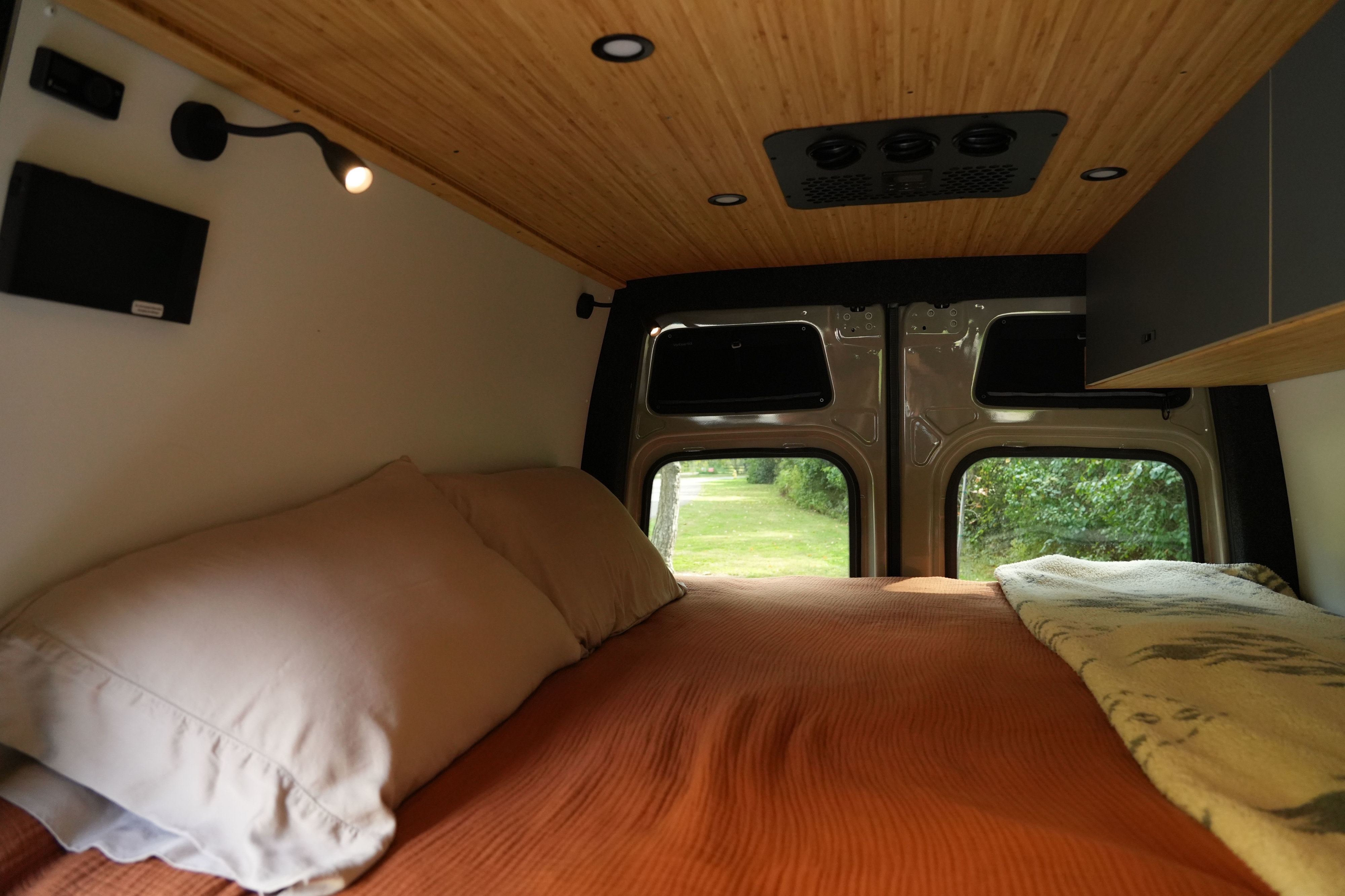Cozy bed in an RV rental, featuring a wooden ceiling, wall-mounted lights, and scenic views from the back windows.. Mercedes Sprinter 2025