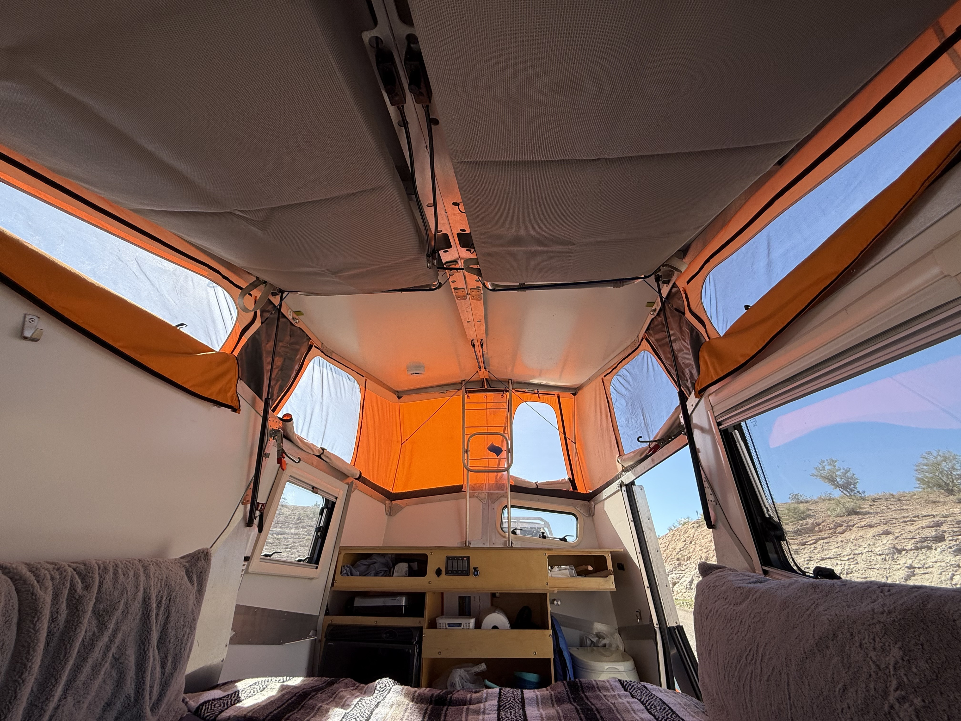 Inside a pop-up rv rental: compact interior with bed, small kitchen, and panoramic windows showing a desert view.. Cricket standard 2013