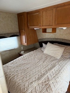 A comfortable RV rental bedroom interior, featuring a queen-sized bed with a cream quilt, overhead wood cabinets, and a window.. Starcraft Autumn Ridge 2009