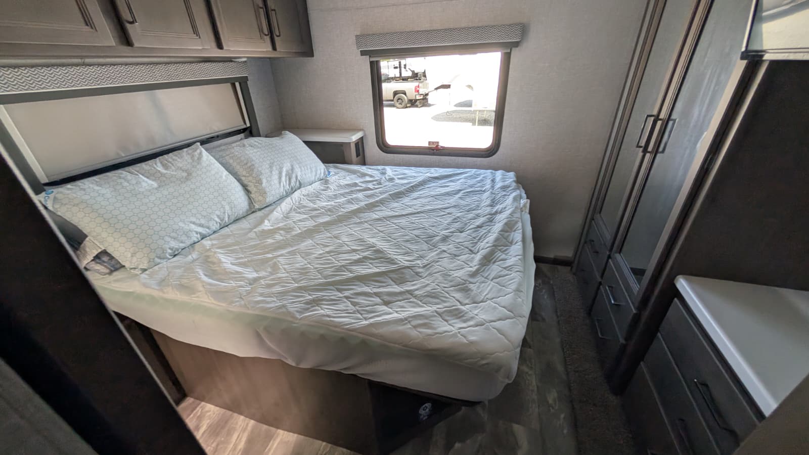 This cozy RV rental bedroom features a comfortable bed with light bedding, overhead cabinets, a window, and built-in storage.. Thor Quantum 2023
