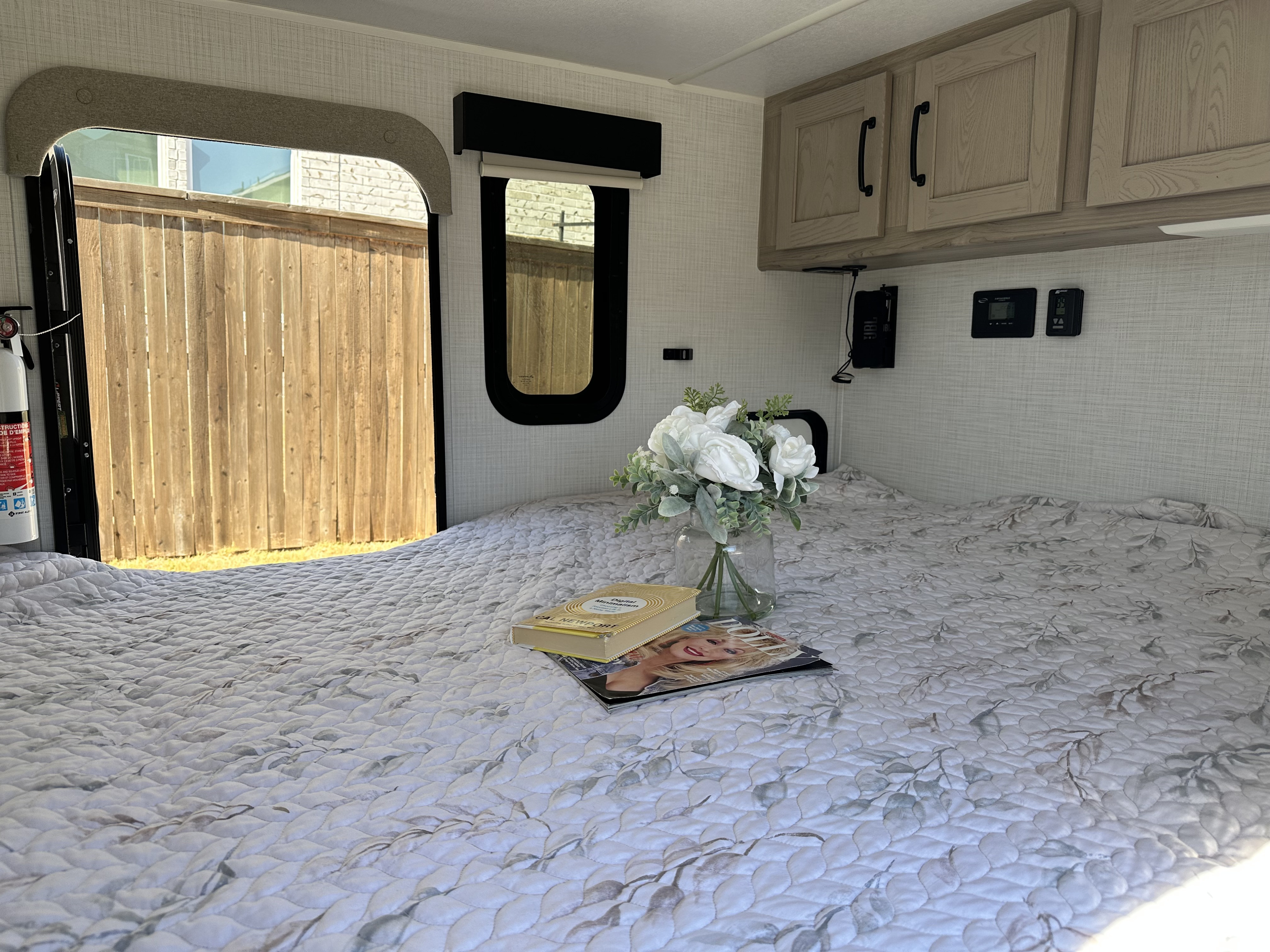 RV rental: Bed with leafy quilt, sunlit windows looking at a fence, overhead cabinets, and a vase of white flowers.. Forest River Flagstaff E-Pro 2024