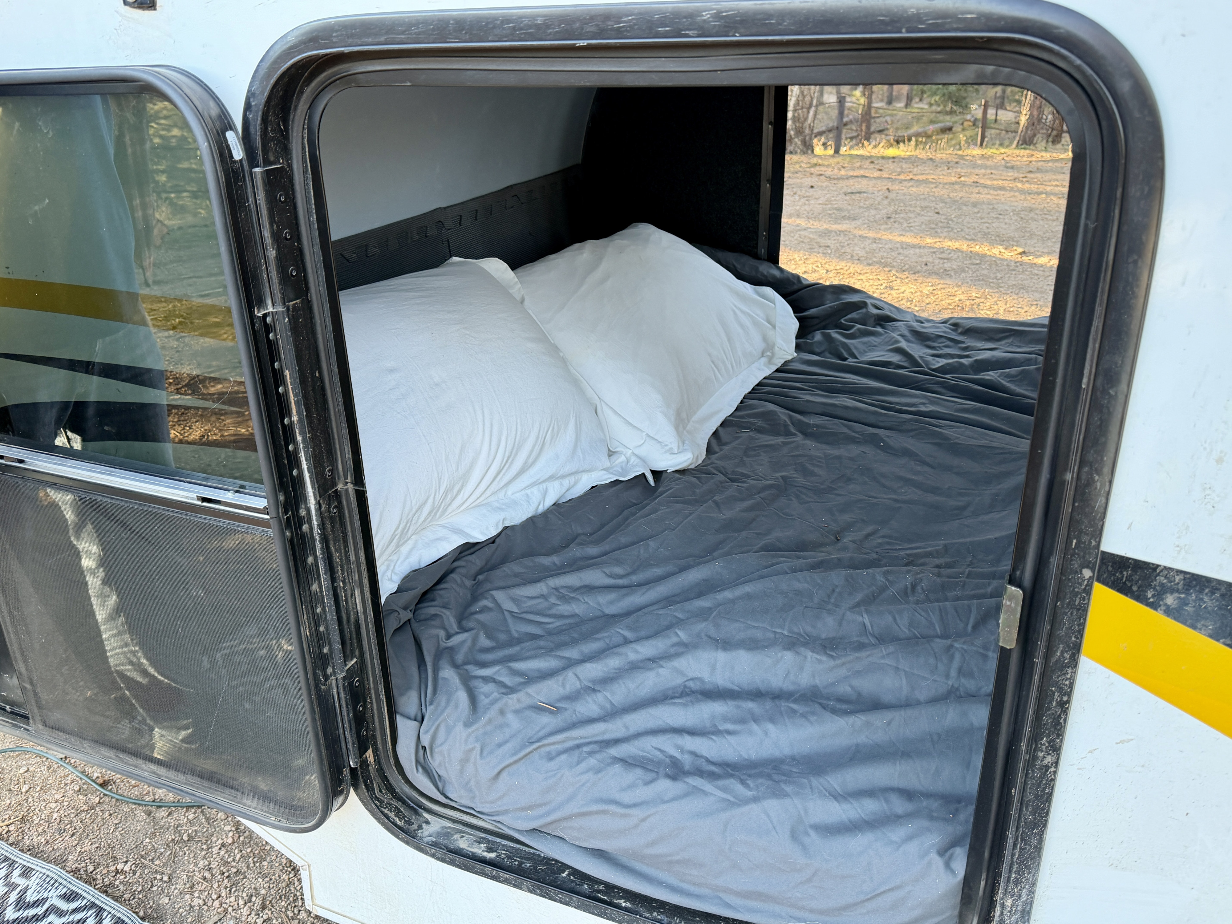 Inside an RV rental's sleeping area, showing a bed with white pillows and gray sheets. Door open to outdoors.. Lil’ Guy RET 2006