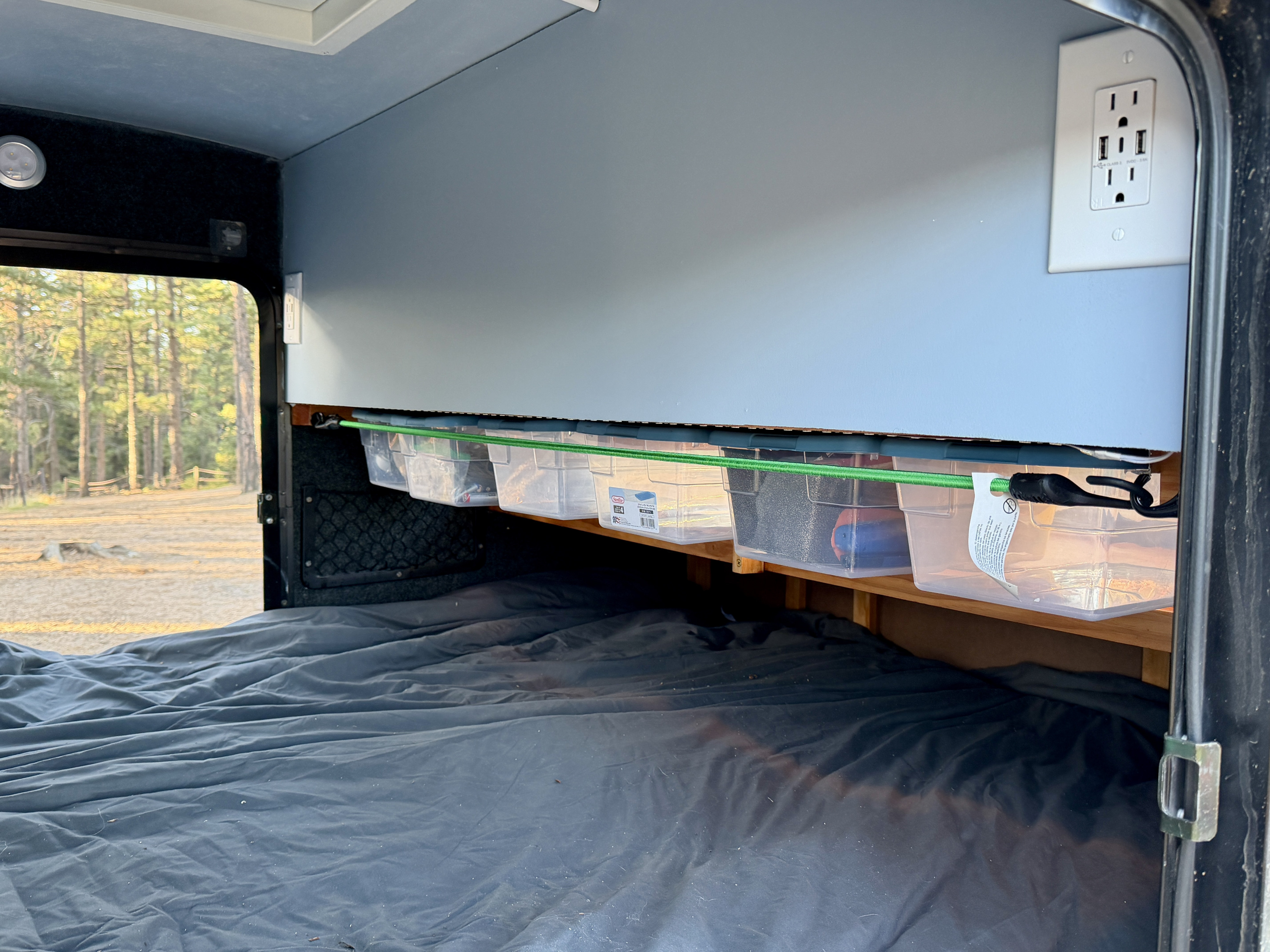 RV rental bedroom with dark bed, overhead storage bins, and power outlet. A green bungee secures bins. Forest outside window.. Lil’ Guy RET 2006