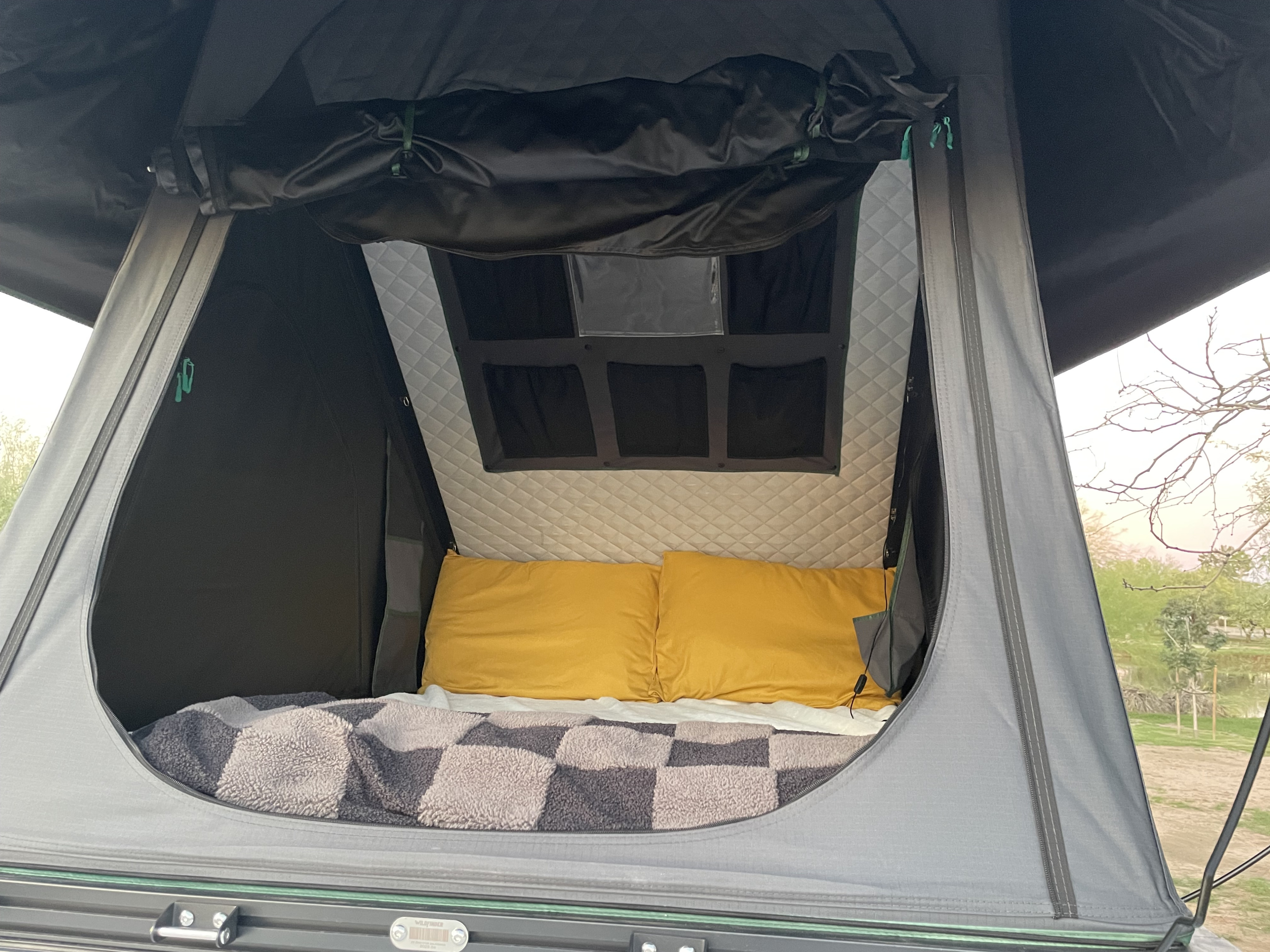 Inside an rv rental rooftop tent: cozy bed with yellow pillows, checkered blanket, quilted walls, and an outdoor view.. Mercedes-Benz Sprinter 2022