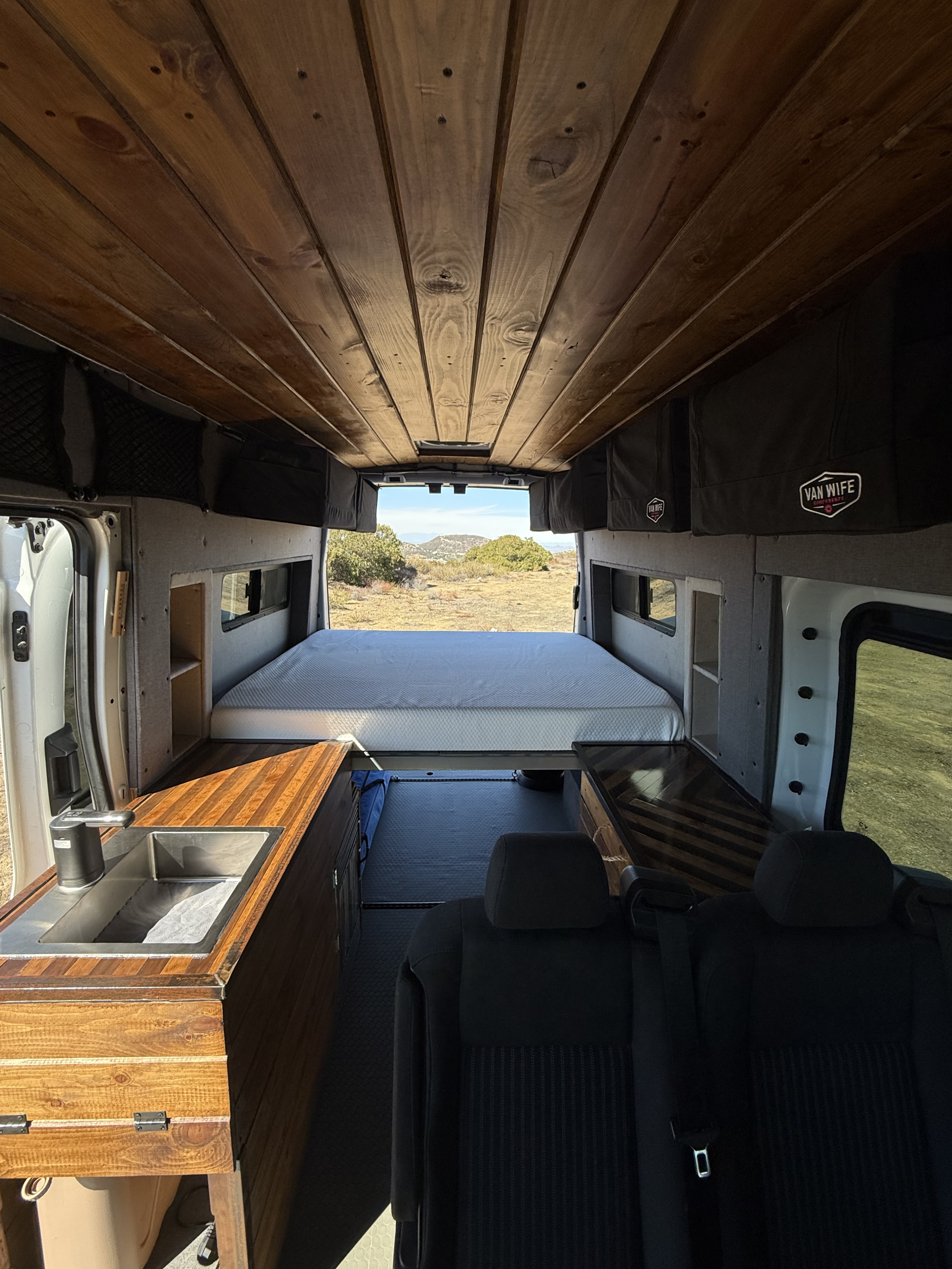 View from inside an rv rental: wood ceiling, kitchen sink, gray bed, and an open rear door revealing a sunny, dry landscape.. Ford T-250 AWD 2022