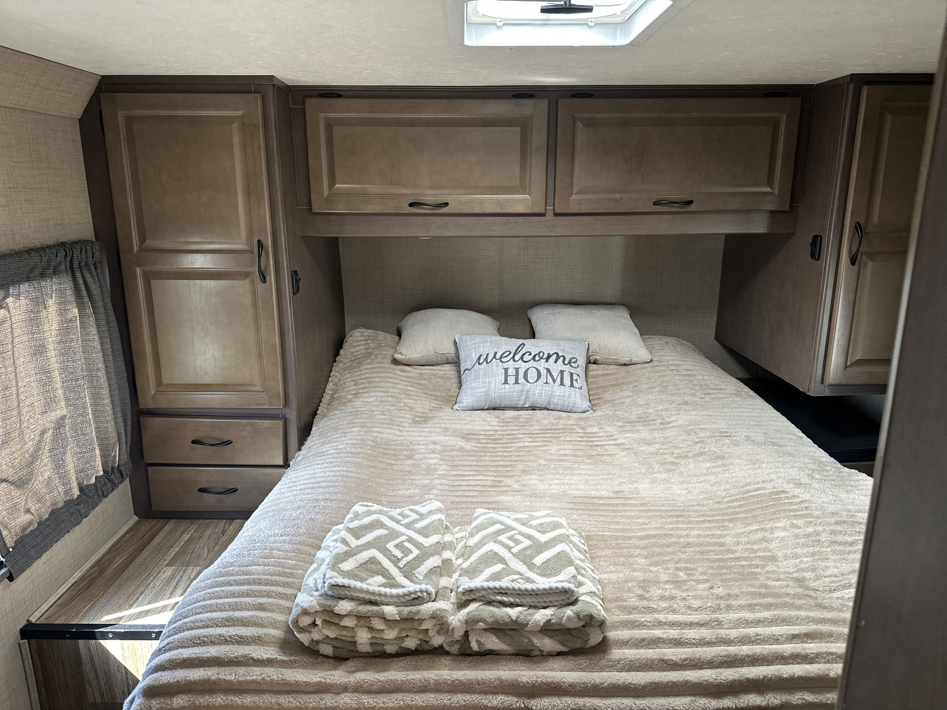 Cozy RV rental bedroom with a comfy bed, "WELCOME HOME" pillow, folded towels, and built-in storage. Ideal travel accommodation.. Winnebago Minnie Winnie 2020
