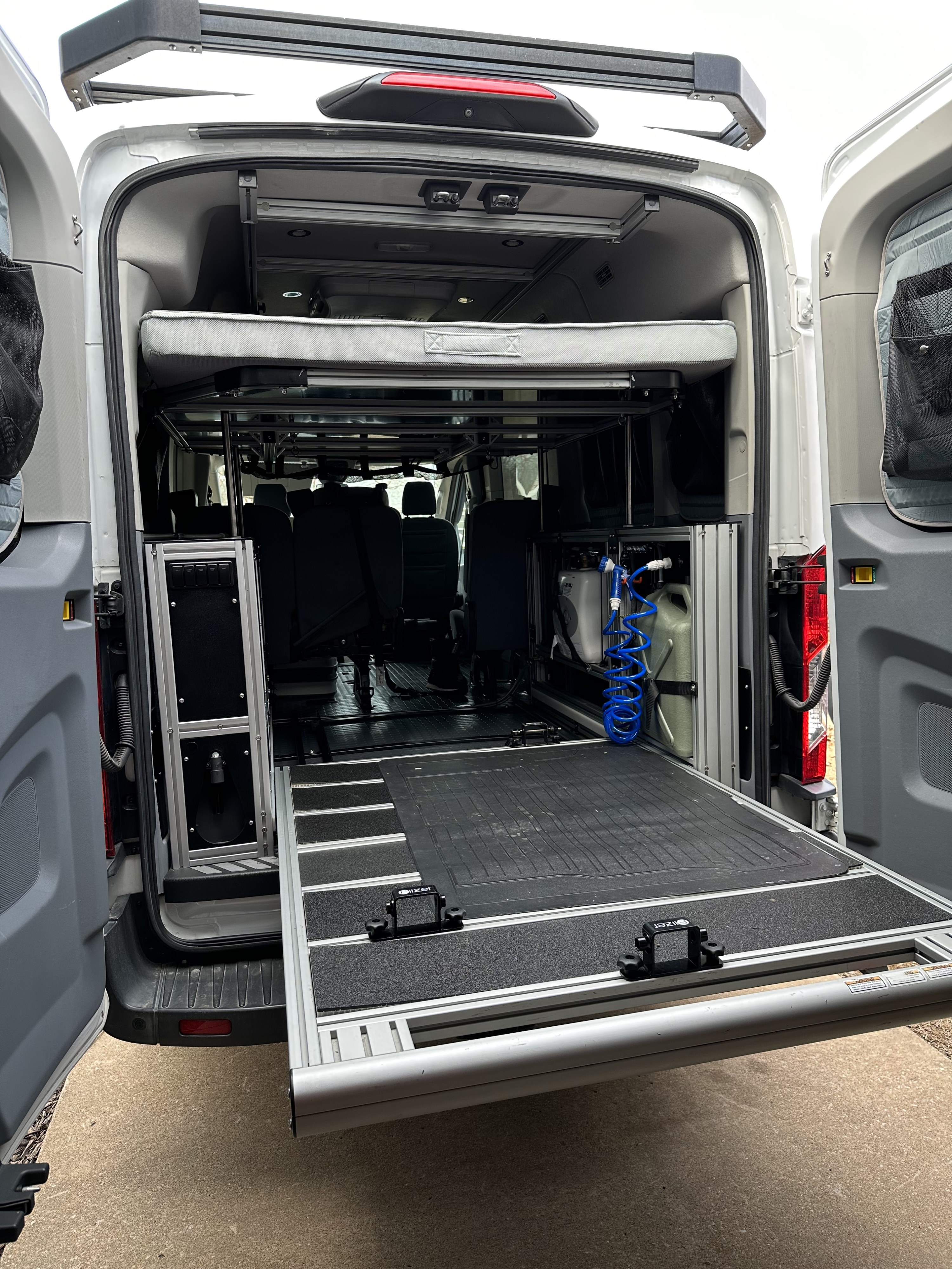 Rear view of a customized RV rental van interior, featuring a pull-out bed, water tanks, and a sliding cargo platform.. Ford Transit 2018