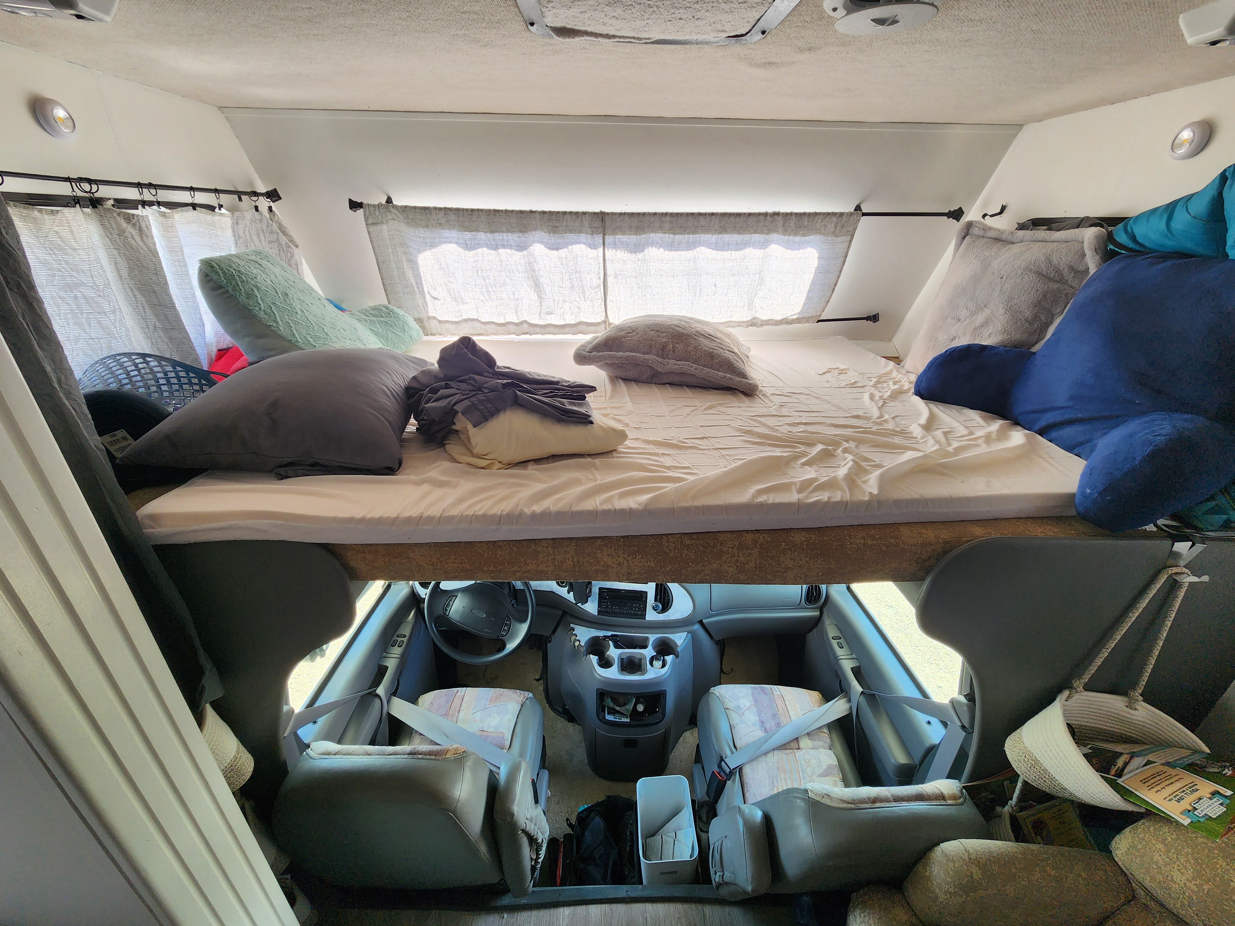 Inside a spacious RV rental: a comfortable overhead bed with pillows and curtains, looking down into the driver's cab and seating.. Jayco Greyhawk 2002