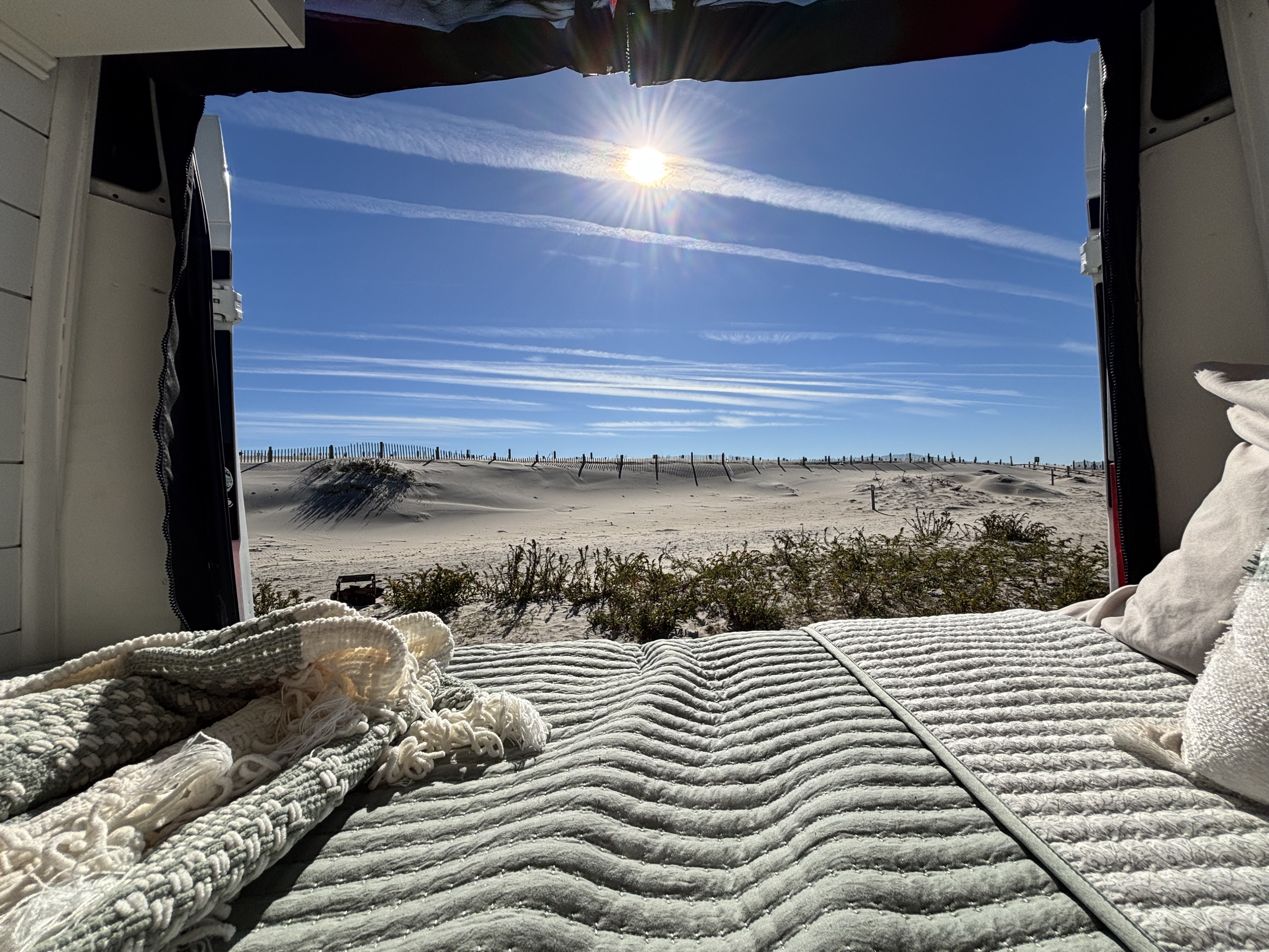 RV rental comfort: a cozy bed with a scenic view of sunny dunes, a beach fence, and a brilliant blue sky.. Ram Ram ProMaster 2500 2019