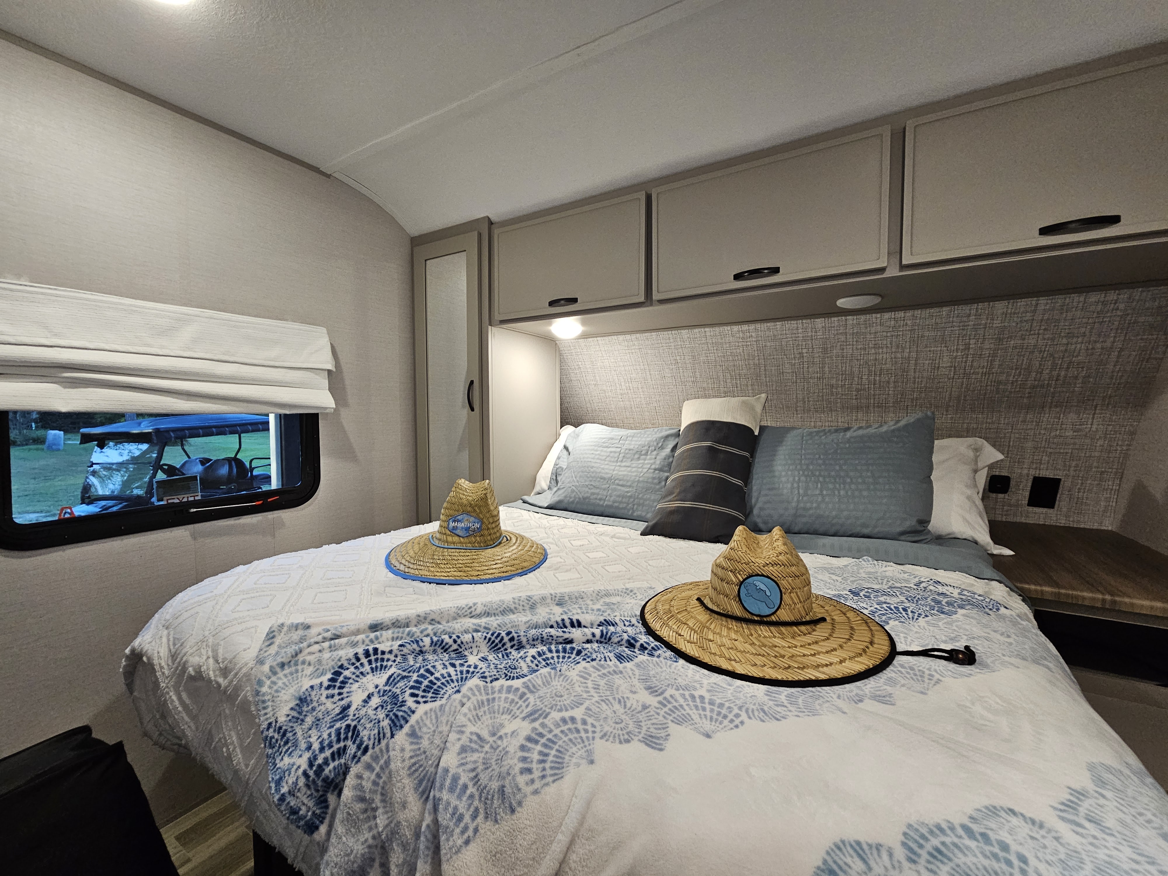 Cozy RV rental bedroom with a comfortable bed, overhead cabinets, and two straw hats. A golf cart is visible through the window.. Forest River Vibe 2024