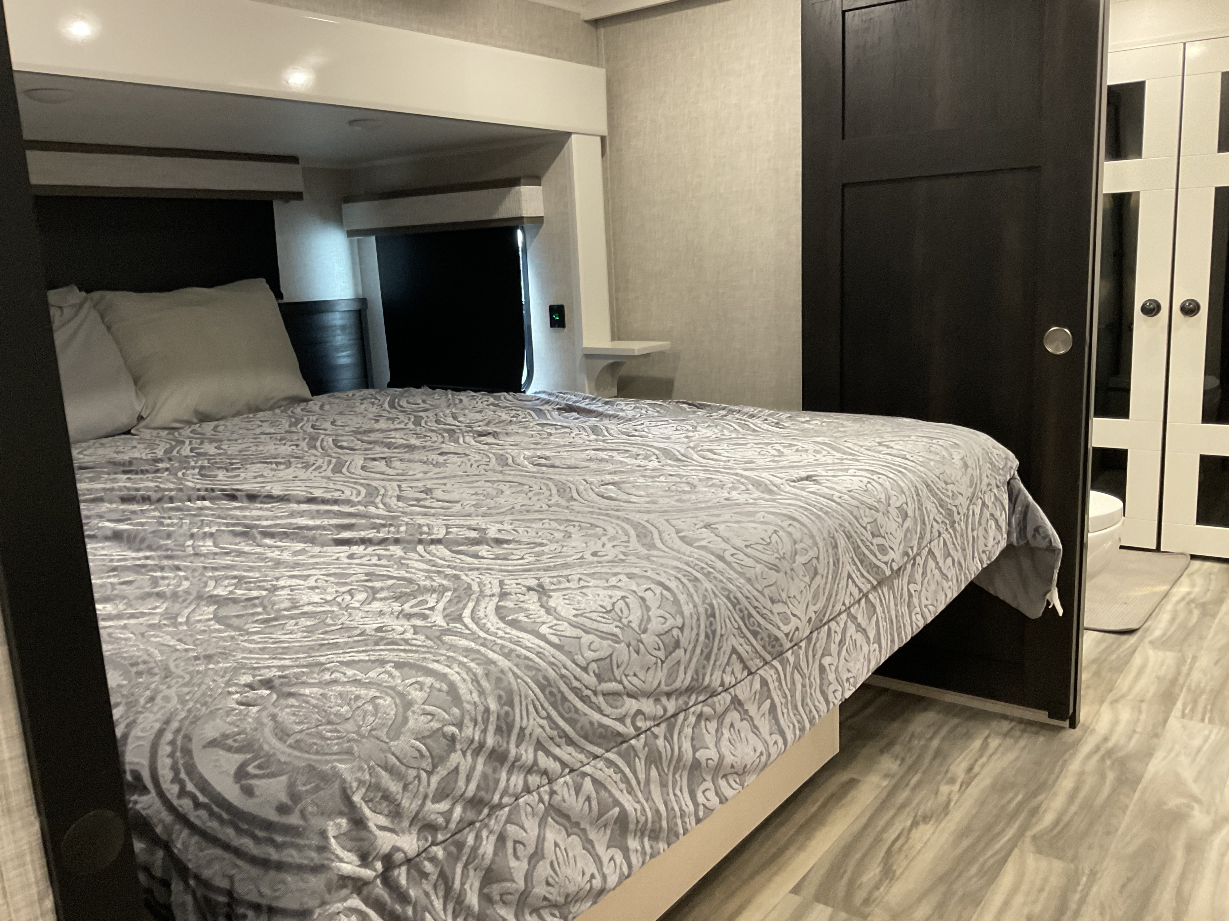 Cozy RV rental bedroom featuring a grey patterned bedspread, dark sliding door, and a glimpse into the compact bathroom.. Jayco Pinnacle 2025