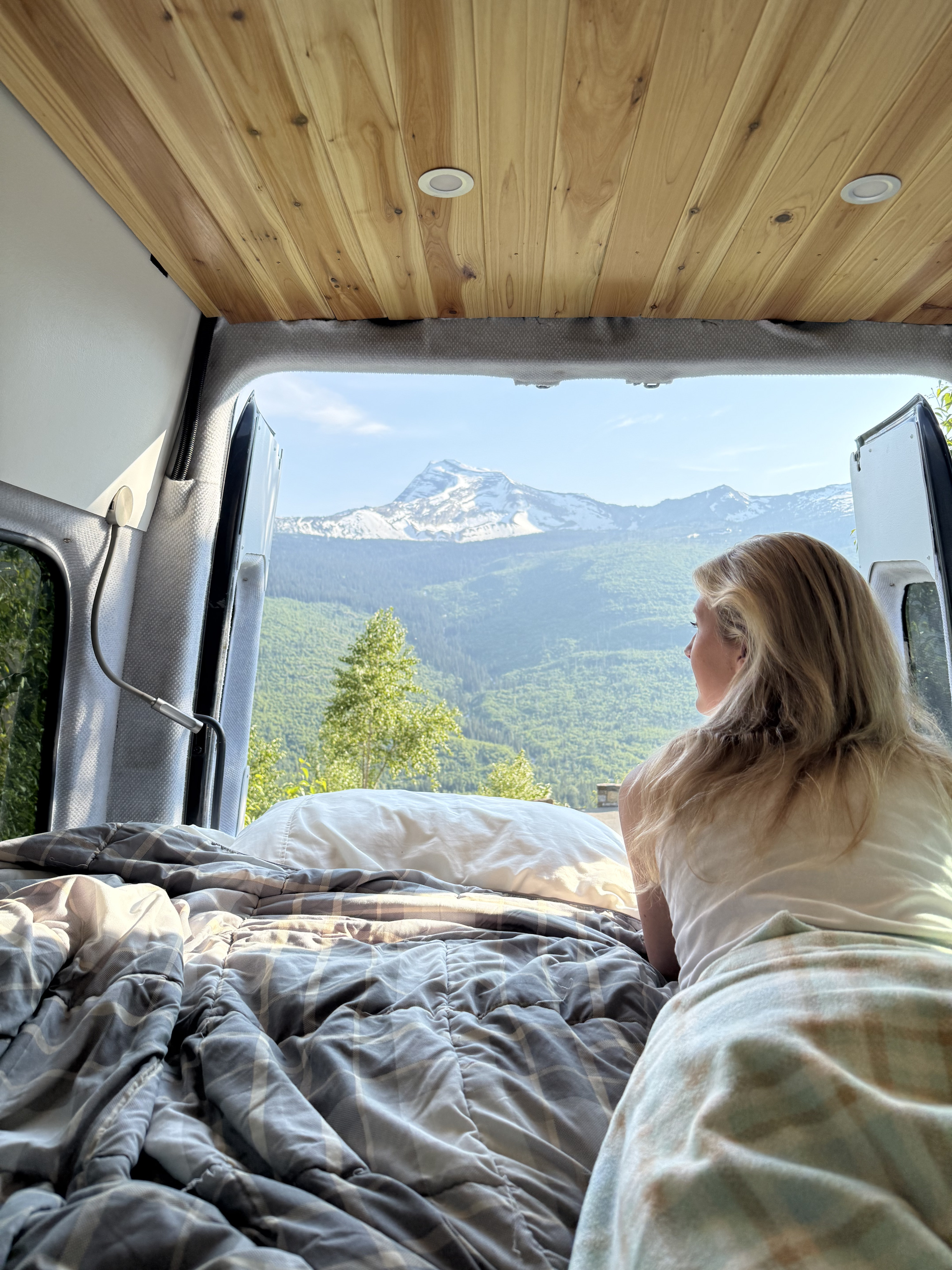 Stunning mountain views from a cozy rv rental bed. A blonde woman relaxes, enjoying the serene van life experience.. Dodge Sprinter 2500 170ext 2008