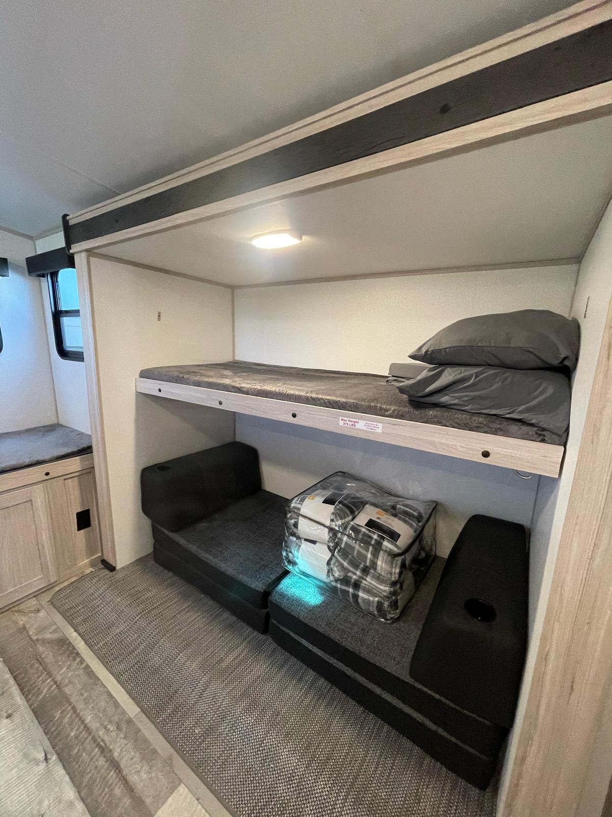 Inside an RV rental: upper bunk bed with mattress/pillows, lower convertible seating/sleeping area, and a side bench with storage.. 2022/SNDC SUDANCE 2022