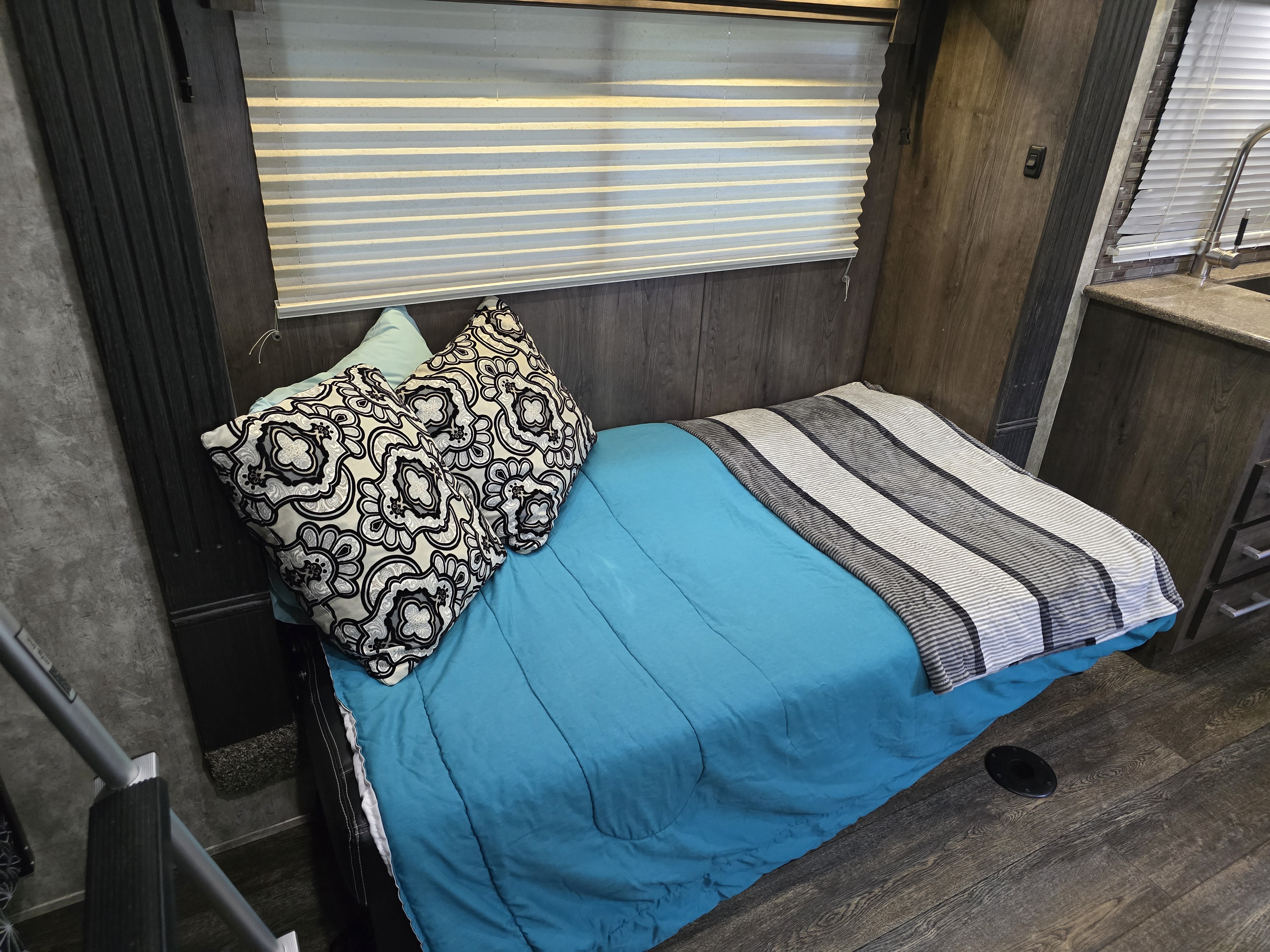 Cozy bed area in an RV rental, with a blue comforter, patterned pillows, wood-paneled walls, and a window with blinds.. Eclipse Recreational Vehicles Attitude 2019