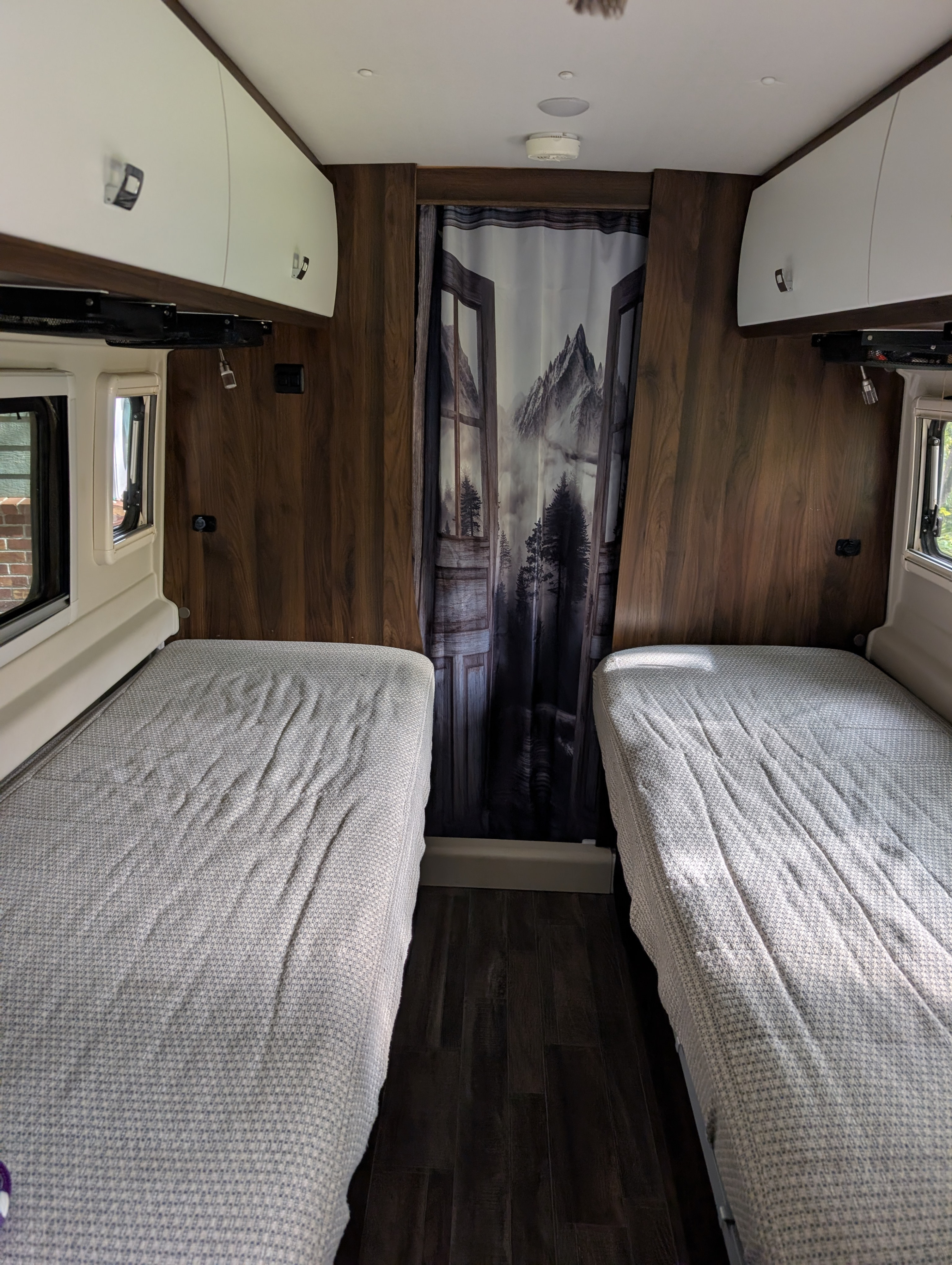 Inside an RV rental: two twin beds with light blankets, overhead storage, and a mountain scene curtain.. Winnebago Travato 2022