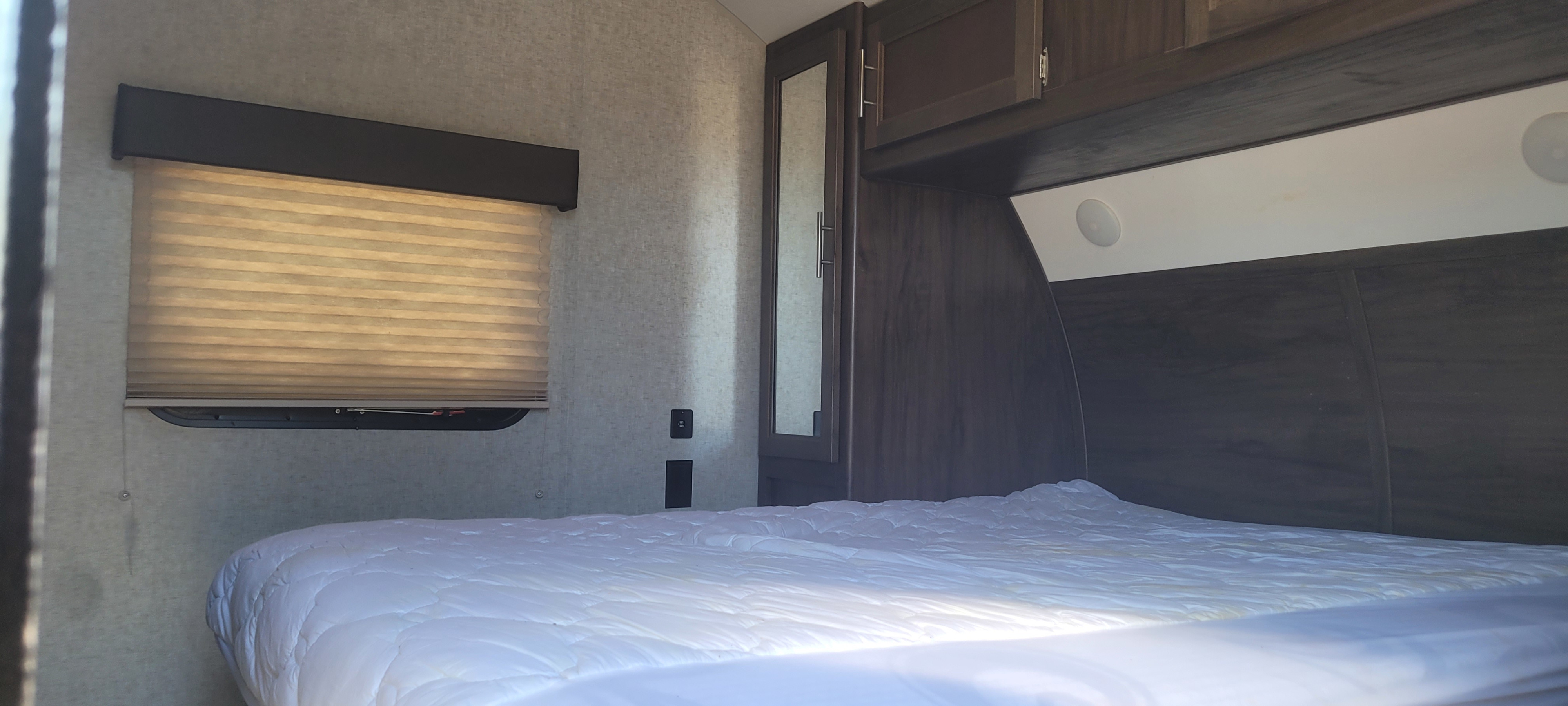 RV rental bedroom featuring a clean white bed, pleated window blind, mirrored cabinet, and overhead storage. Compact sleeping area.. Forest River Cherokee 2018