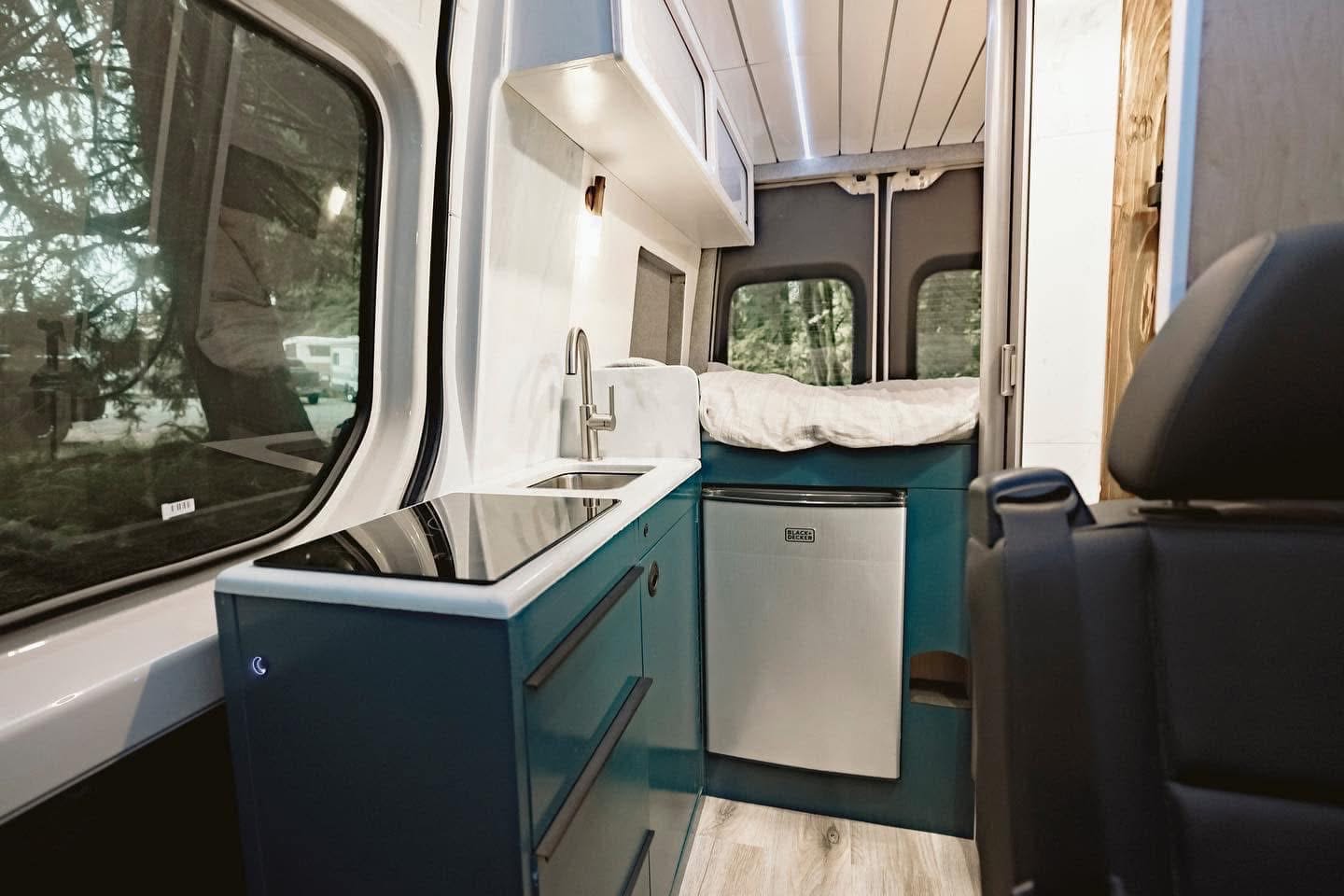 Inside a modern rv rental: compact camper van kitchen with sink, induction cooktop, fridge, and cozy bed in the rear.. Mercedes Benz Sprinter 2020