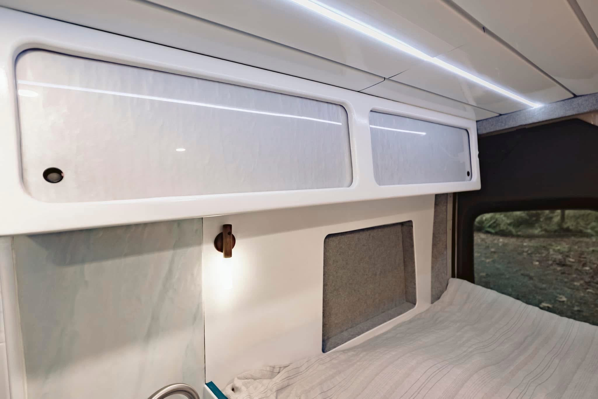 Stylish RV rental interior features white overhead cabinets, sleek LED lighting, a small wooden lamp, and a cozy bed by a window.. Mercedes Benz Sprinter 2020
