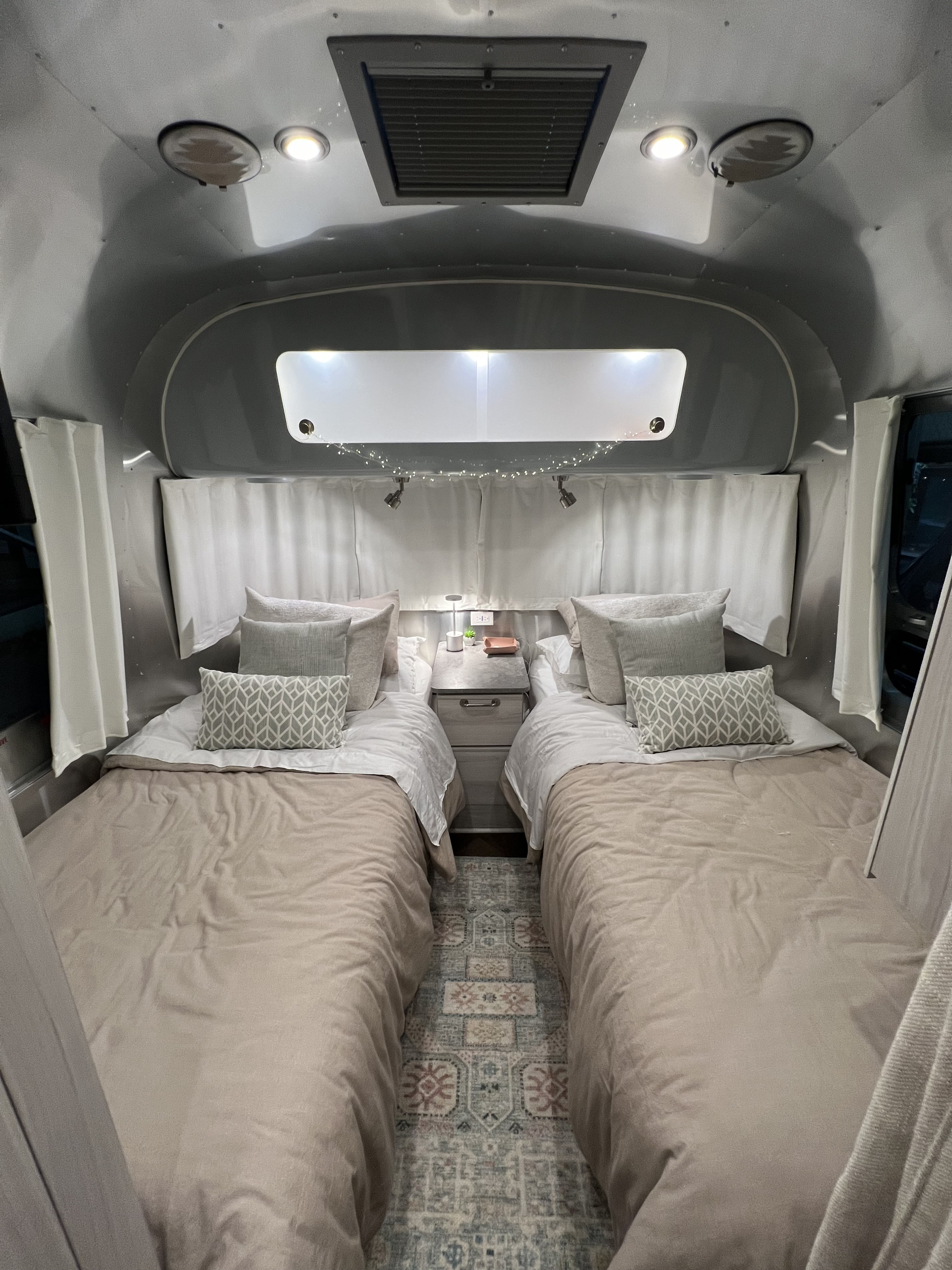 Cozy RV rental bedroom with twin beds, a nightstand, soft lighting, and patterned rug, ideal for a comfortable road trip.. Airstream International 2023
