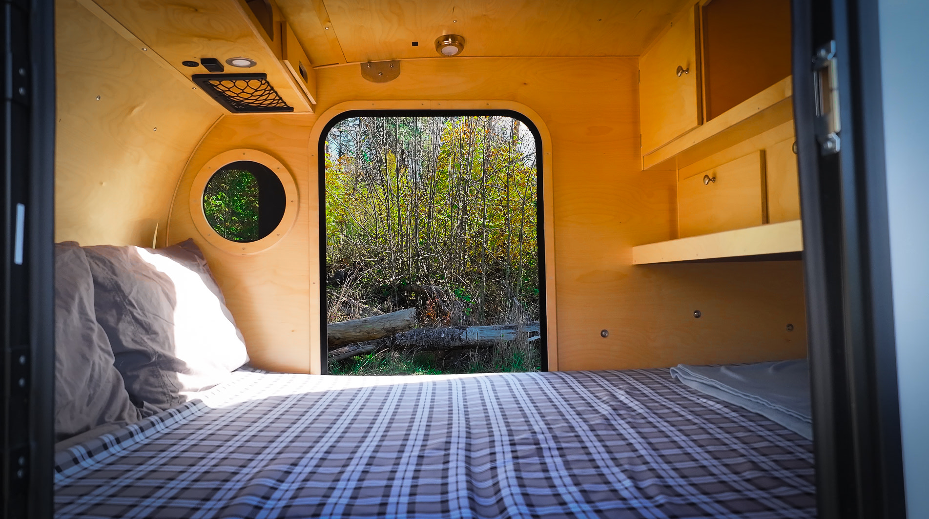 Step into this cozy RV rental's wood interior: a comfy bed, round window, and shelves, with nature outside.. Teardrops NW Scout 2024