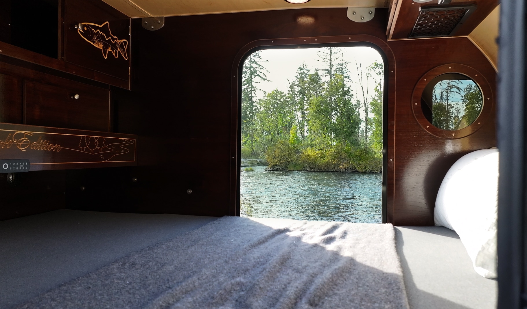 An RV rental's cozy bed looking out onto a peaceful river and forest, offering a tranquil escape into nature.. Teardrops NW Ranger 2025