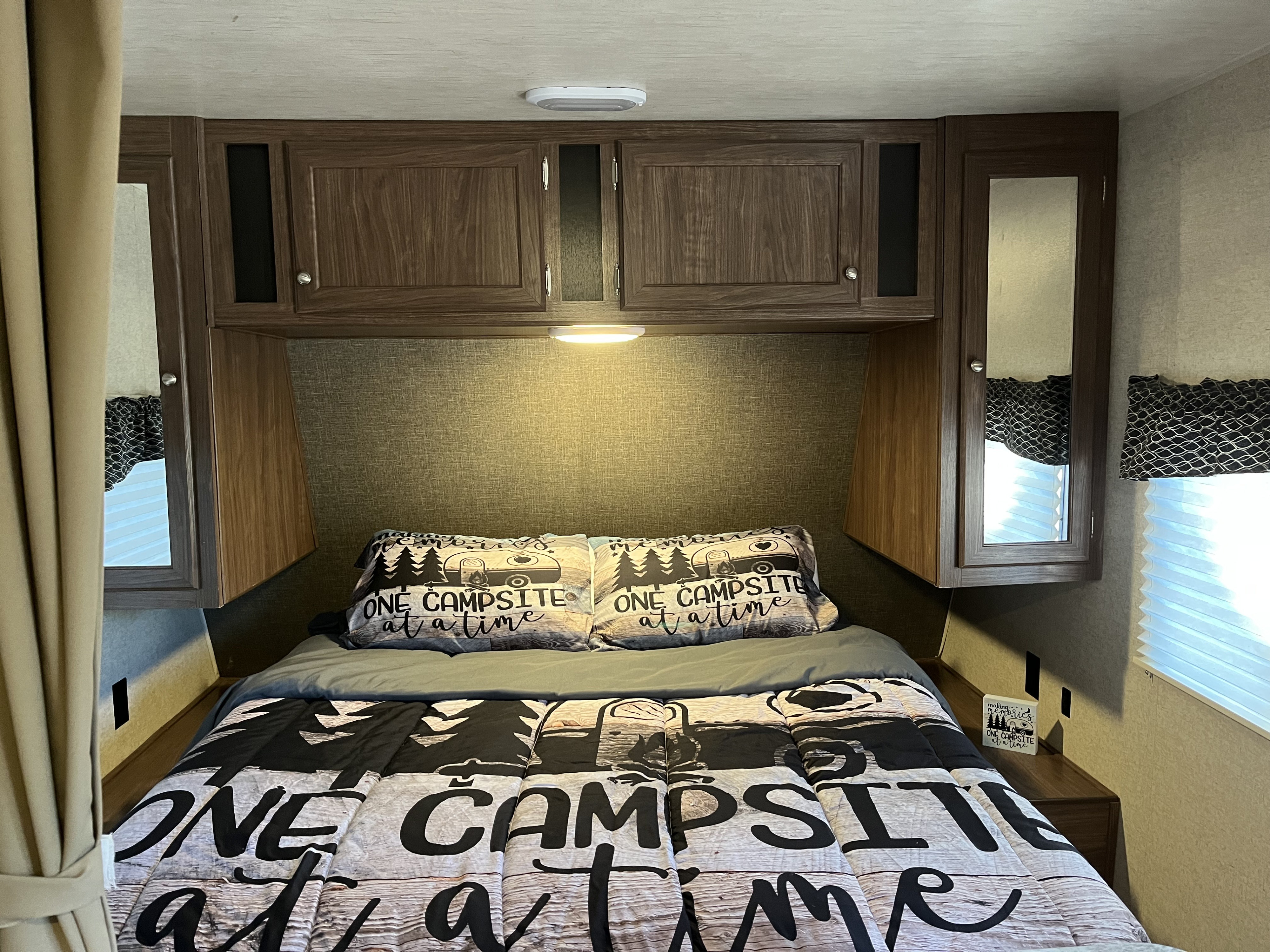 Cozy RV rental bedroom featuring "One Campsite at a Time" bedding, overhead cabinets, and a window for comfortable travel.. Heartland RVs Pioneer 2018