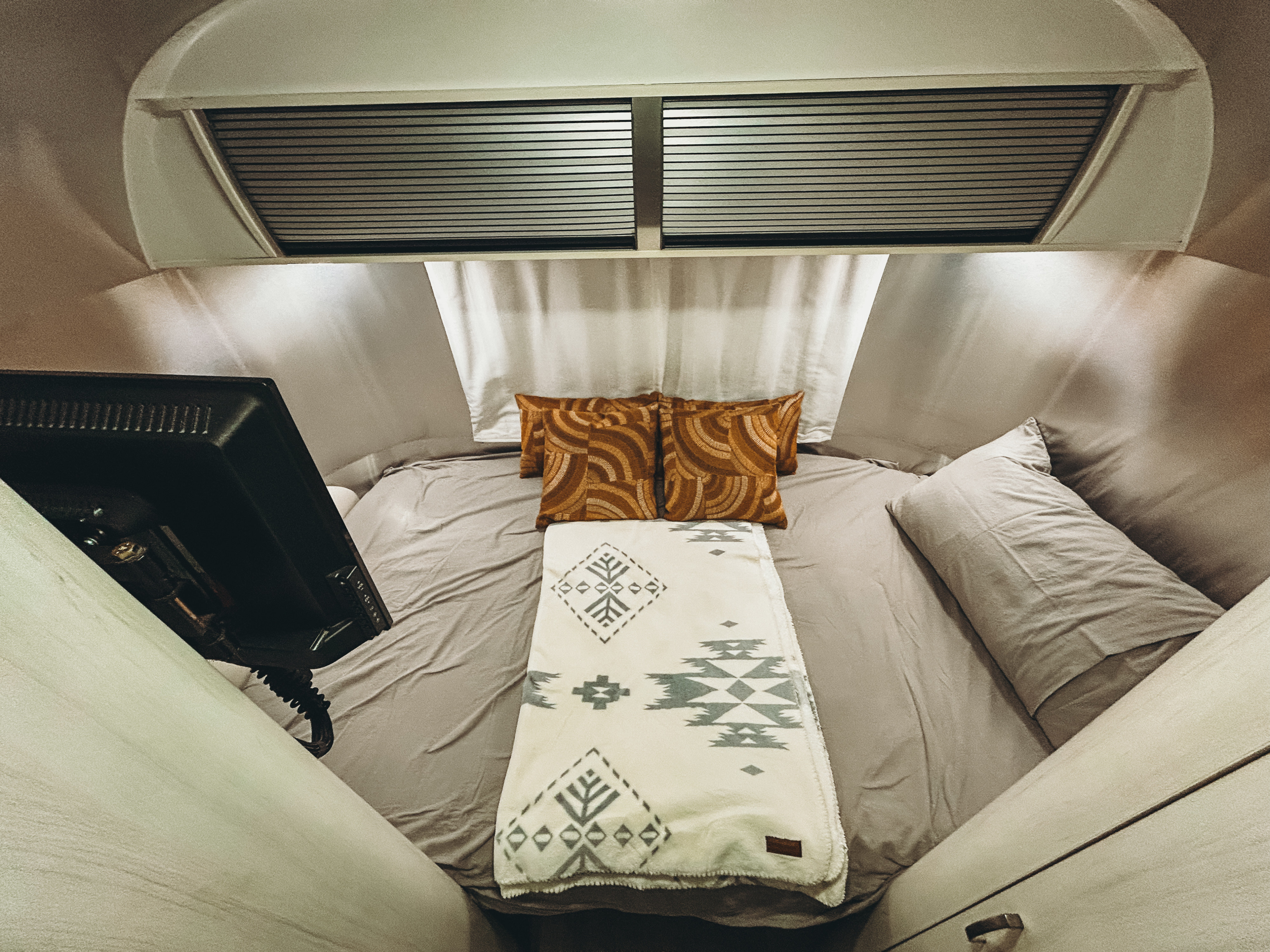 Inside a comfy RV rental bedroom: grey bed, orange patterned pillows, geometric blanket, and TV. Ideal for road trip nights.. Airstream Sport 2016