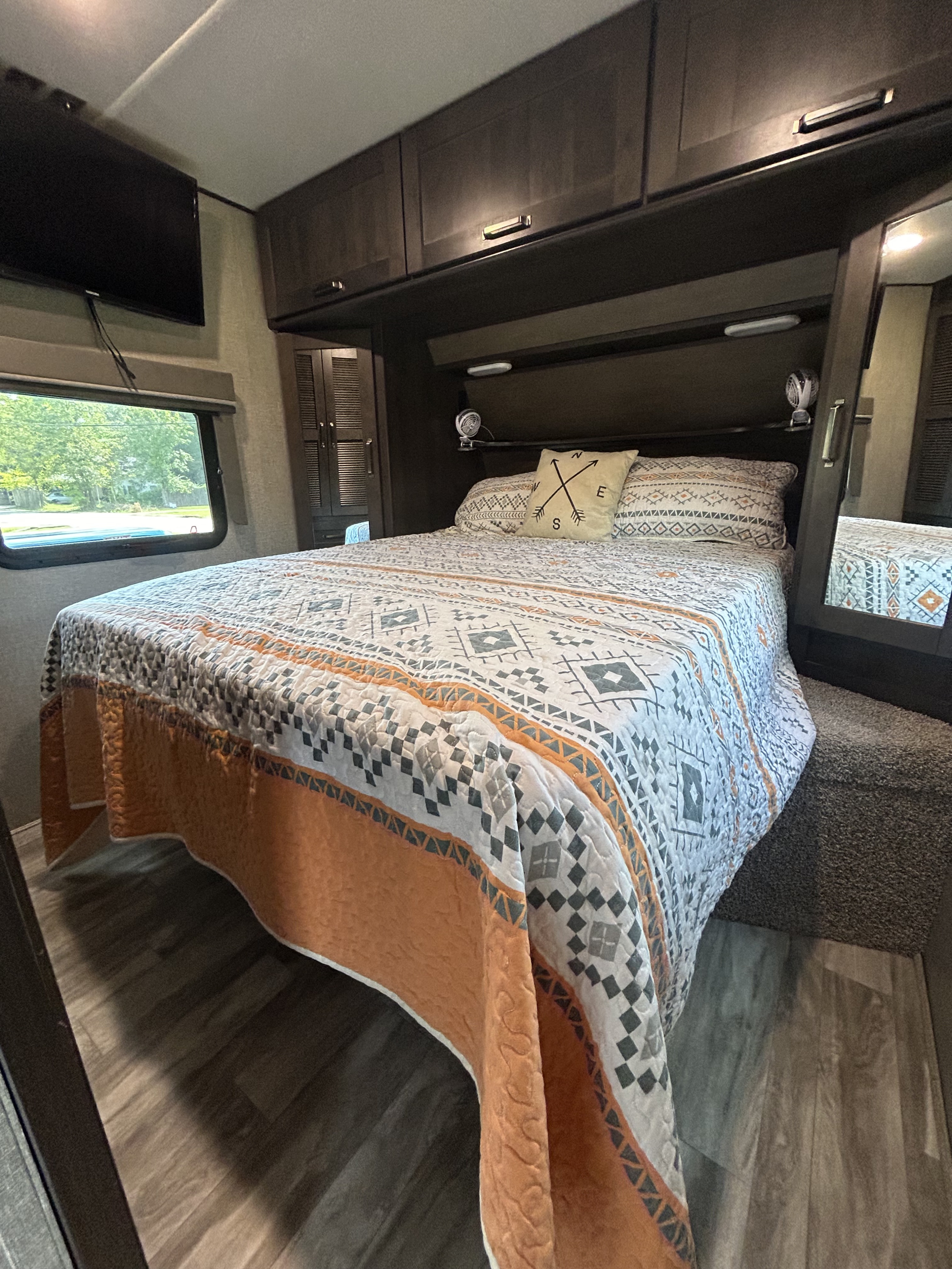 This RV rental features a comfortable bedroom with a patterned bed, dark wood storage, TV, and window view.. Grand Design Reflection 2021