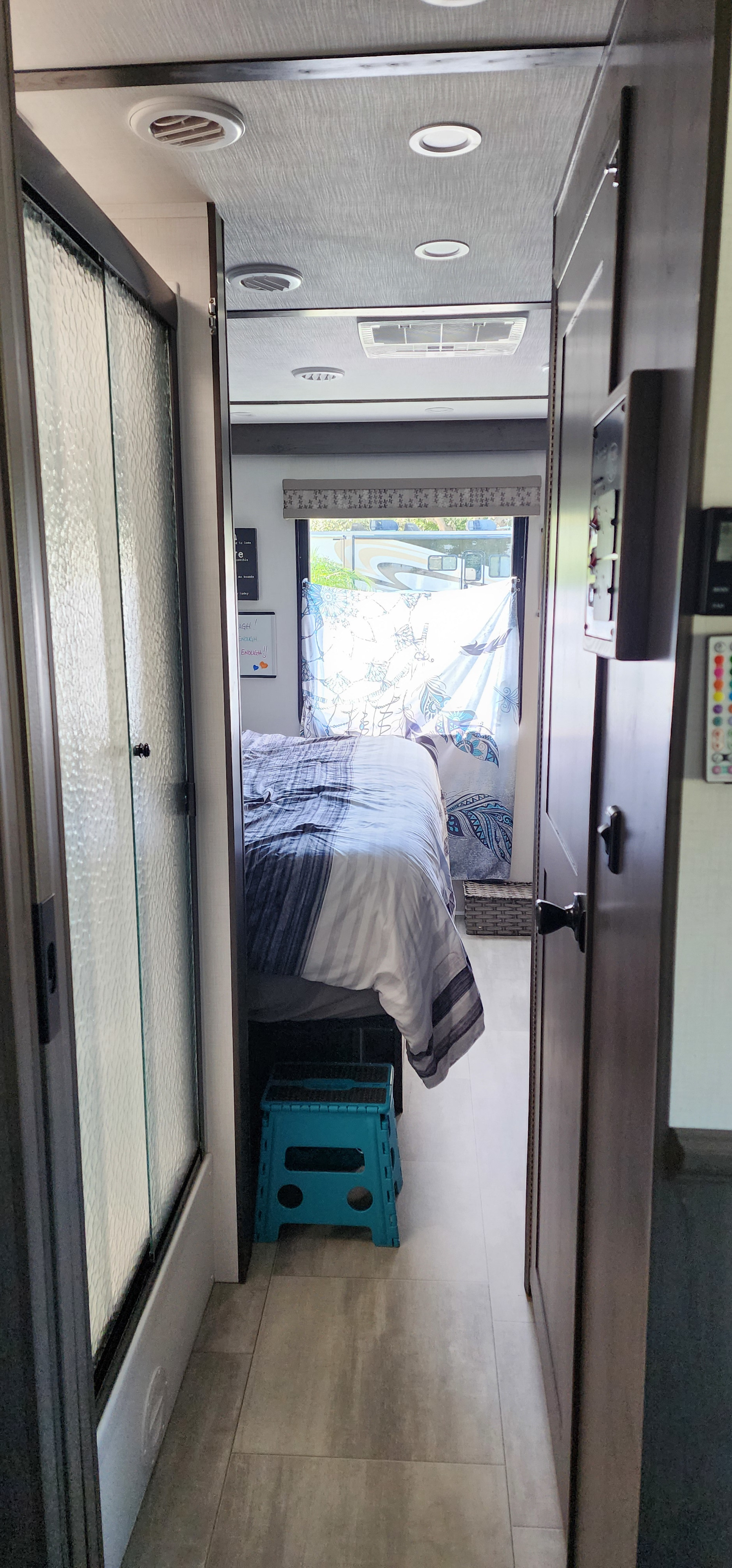 Inside an RV rental: a compact hallway leading to the bedroom. Shower stall left, dark wood door right, bed ahead by window.. Forest River FR3 2024